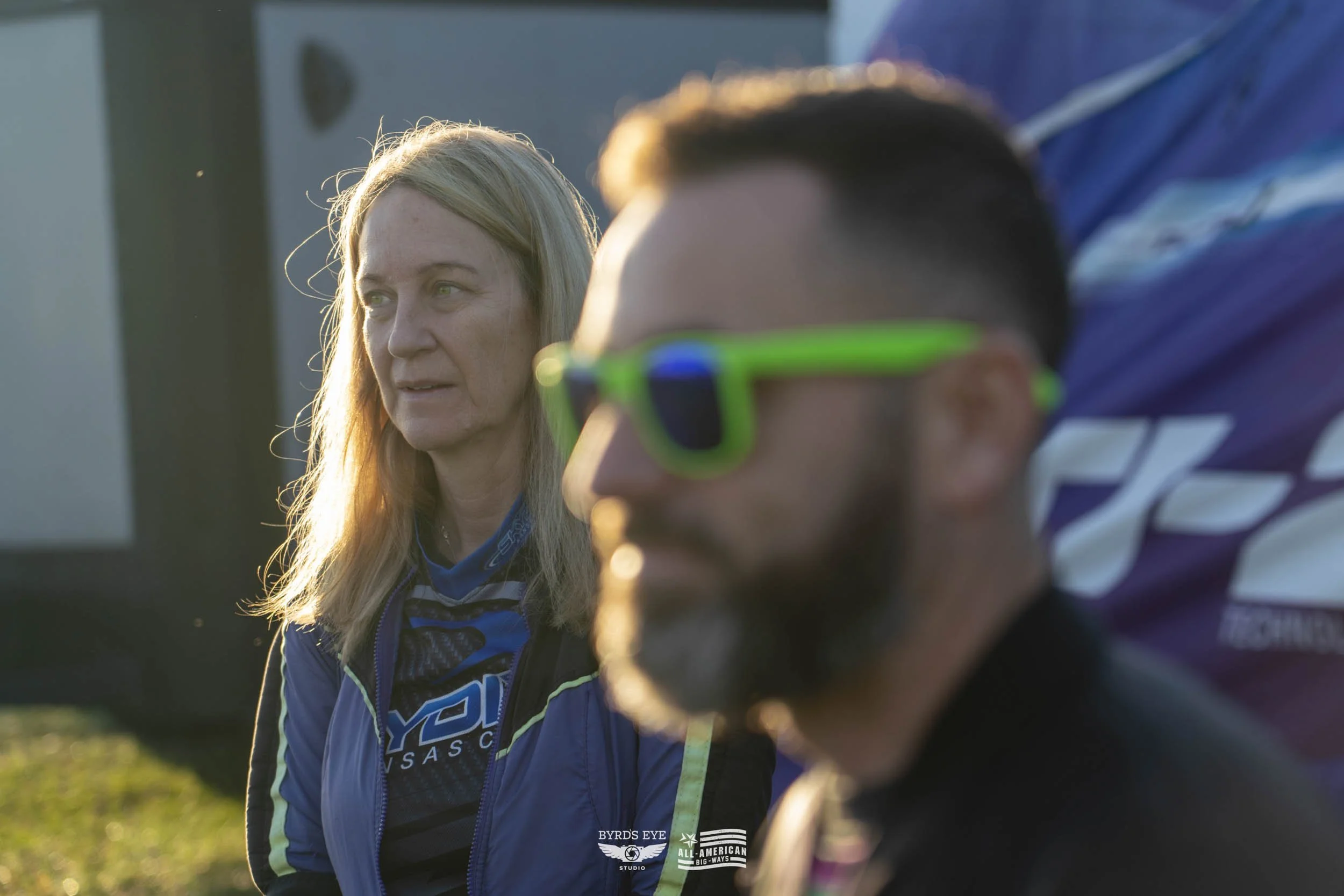 A woman and a man sitting outdoors, with the woman looking to the side and the man wearing bright green sunglasses, during sunset.