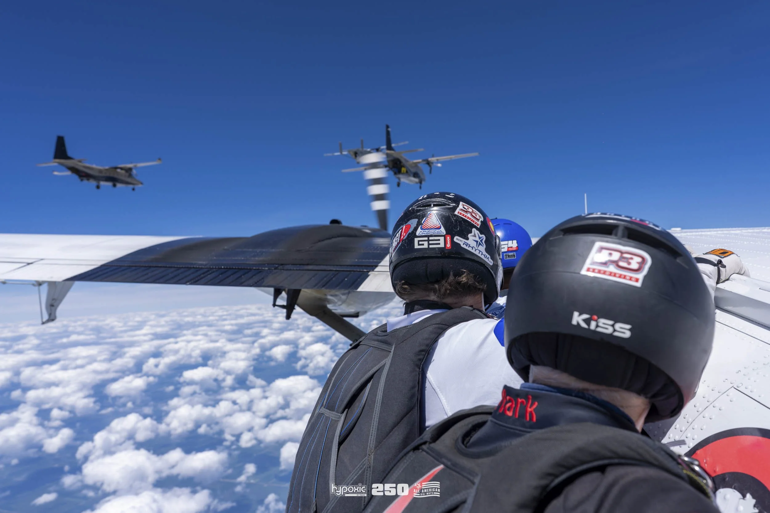 Two pilots in helmets and flight suits on the wing of a small aircraft flying high above the clouds, with two military planes flying in formation in the blue sky behind them.
