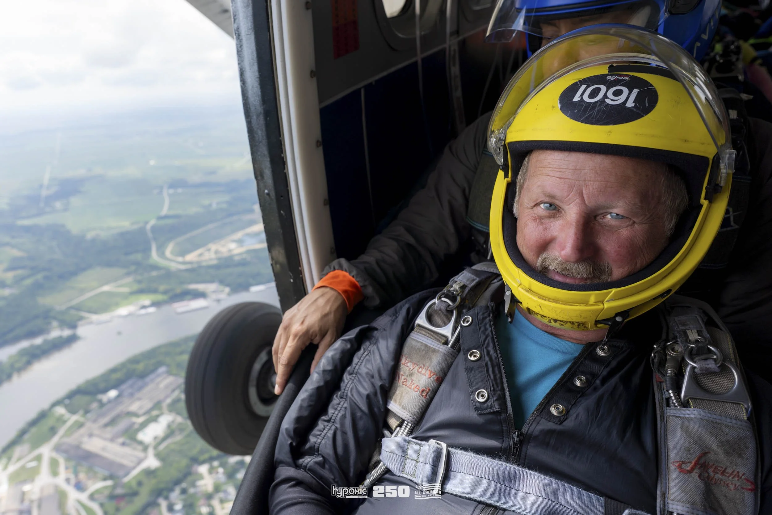 A man with a yellow pilot helmet and jumpsuit, sitting inside a helicopter with another person behind him, preparing for a skydiving jump over a landscape with water, roads, and buildings.