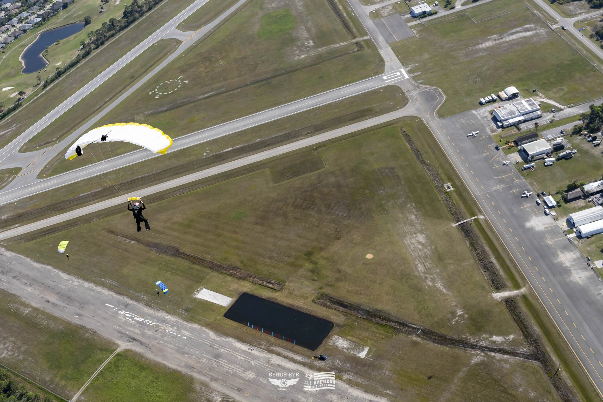 A person parachuting down near a small pond on an airfield surrounded by grass fields, airport roads, parking lots, small buildings, and aircraft.