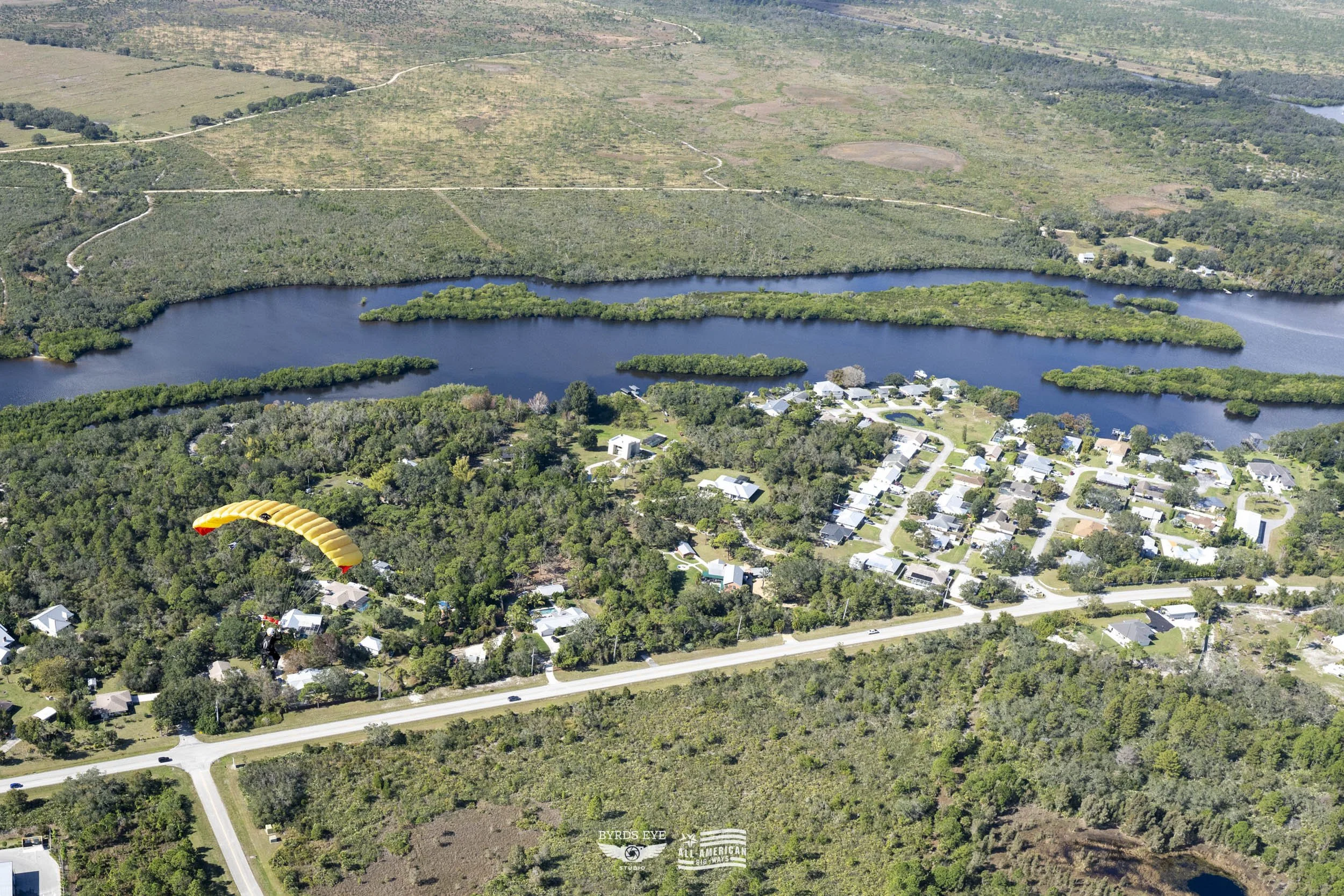 An aerial view of a small town with a lake, surrounded by trees and green landscape, with a person paragliding over the trees.