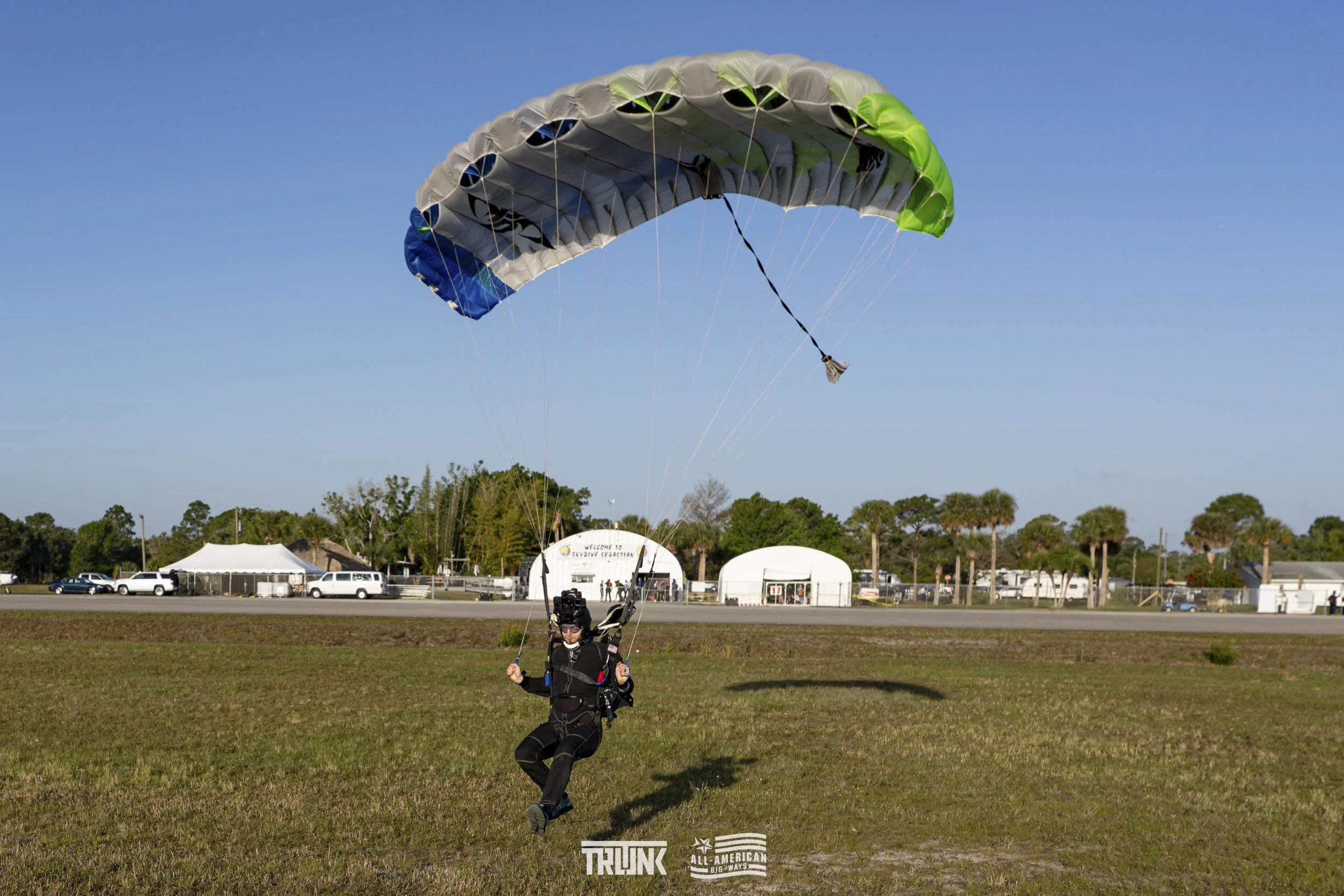 Person landing after paragliding flight on grass field with tents and trees in background under clear blue sky.