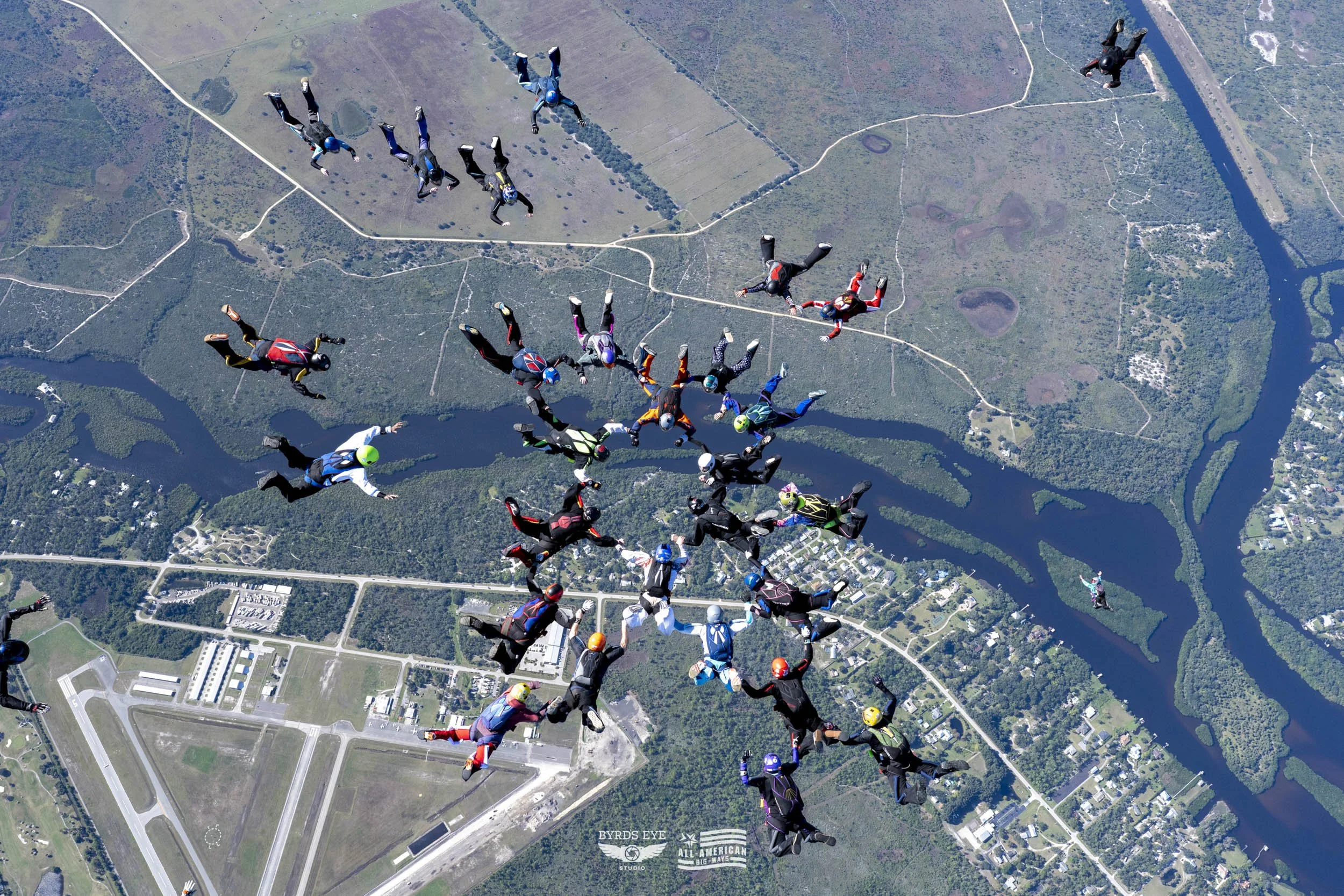 Group of skydivers in freefall over a landscape with lakes, trees, roads, and an airfield.