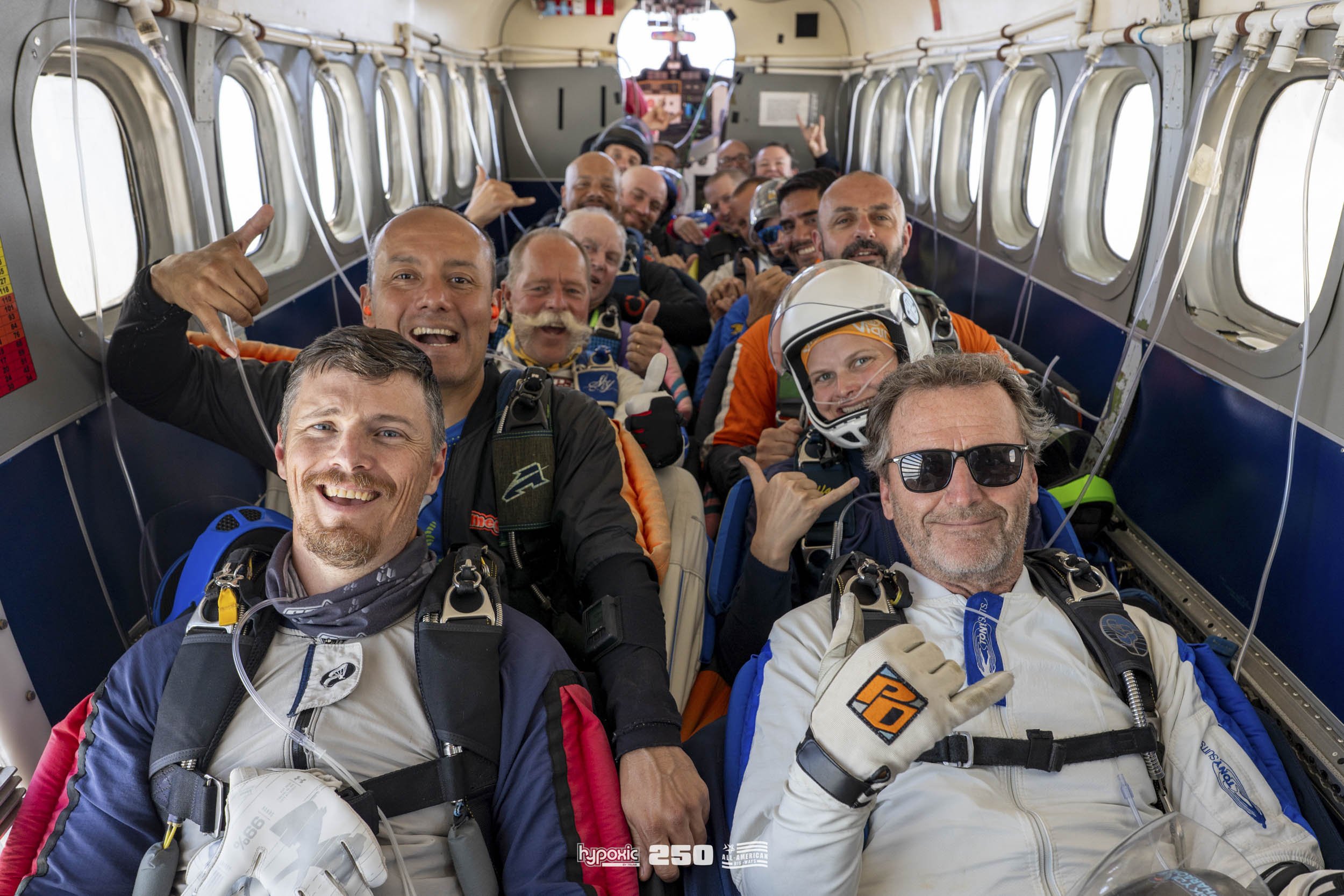 A group of skydivers sitting in the interior of an aircraft, smiling and making hand gestures, wearing jumpsuits and gear, preparing for a jump.