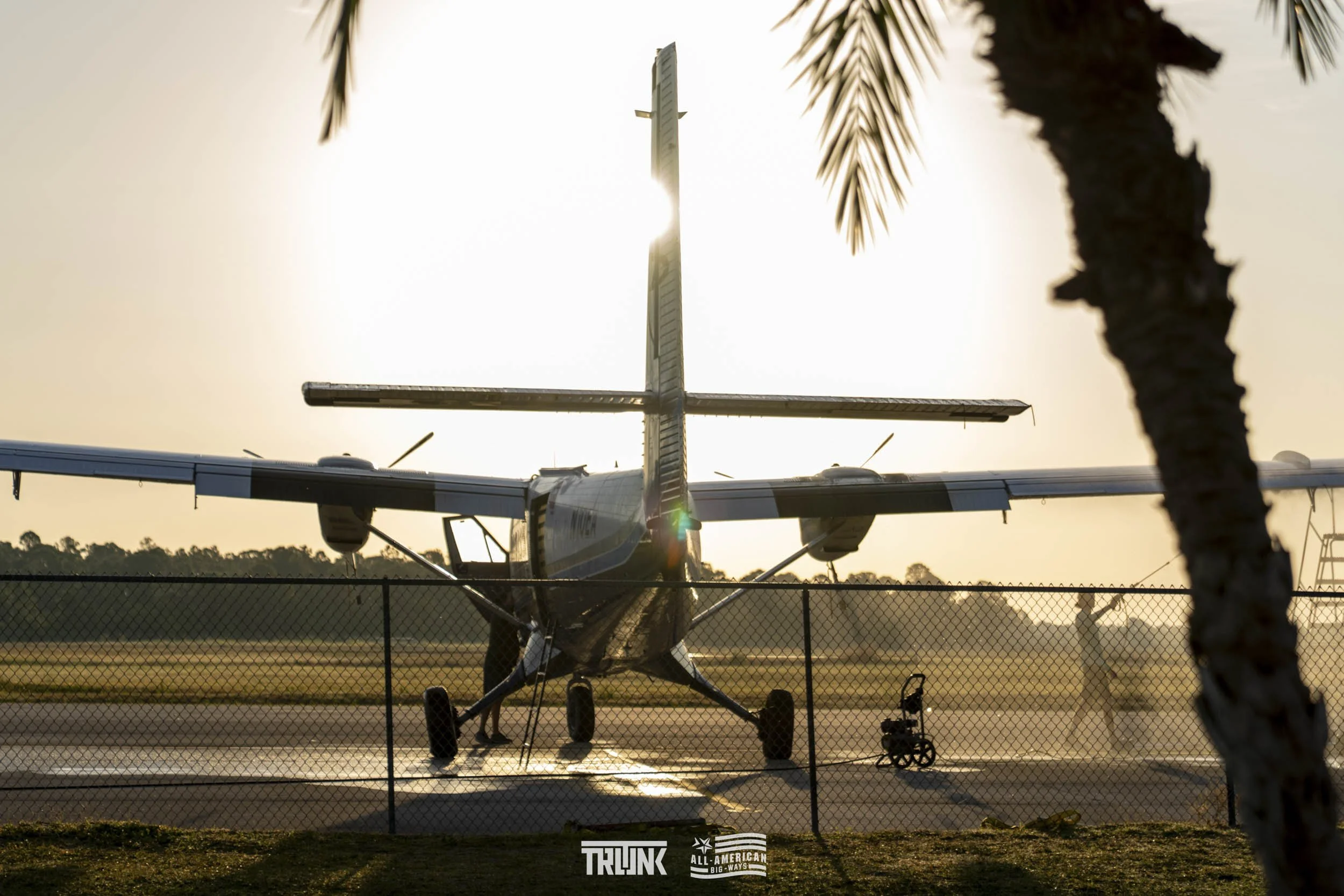 Small airplane on a tarmac with a chain-link fence in the foreground, palm tree on the right, and a person walking away in the background during sunset.