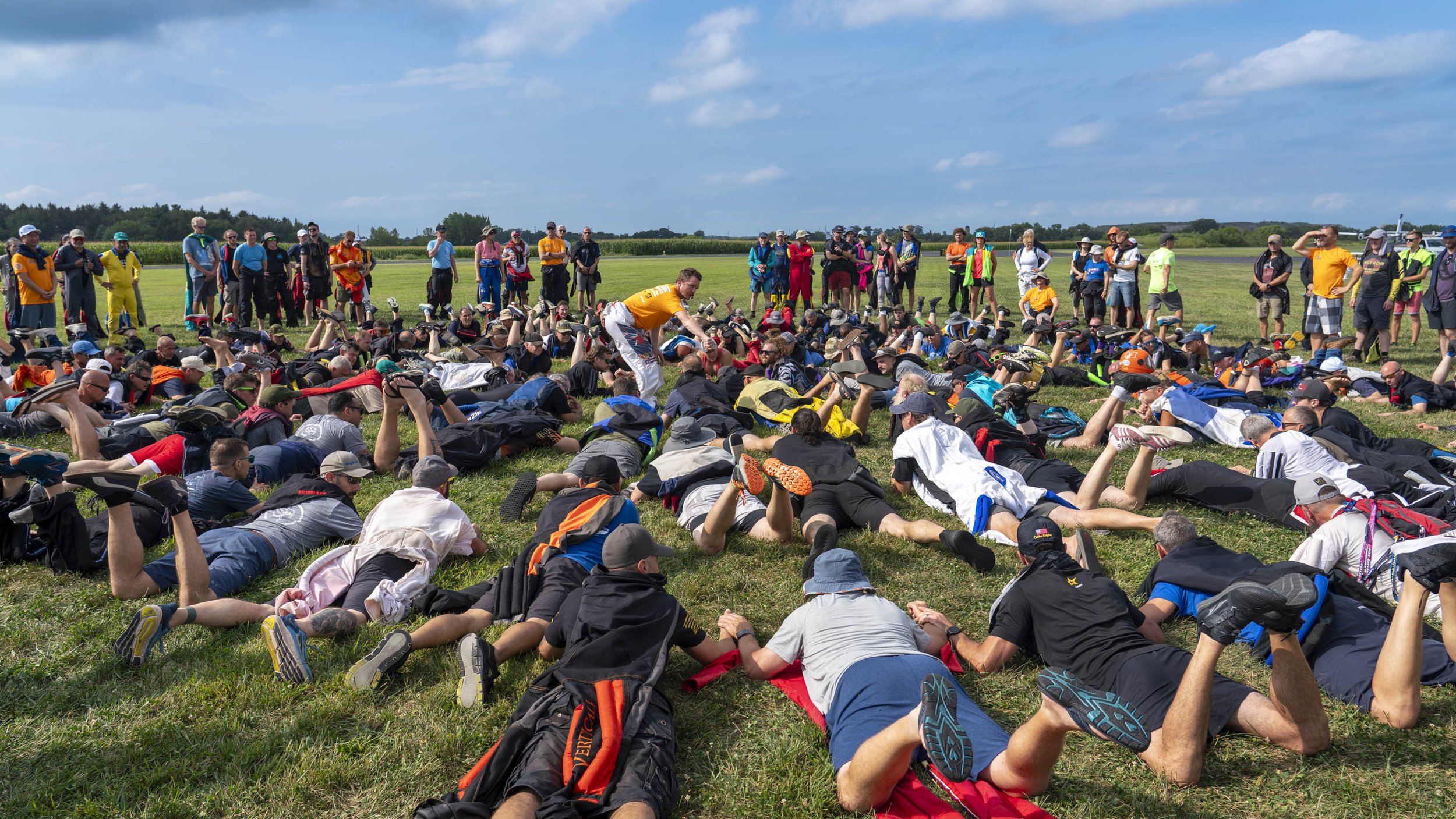 A large group of people participating in an exercise or fitness activity outdoors on a grassy field under a partly cloudy sky, with others observing.