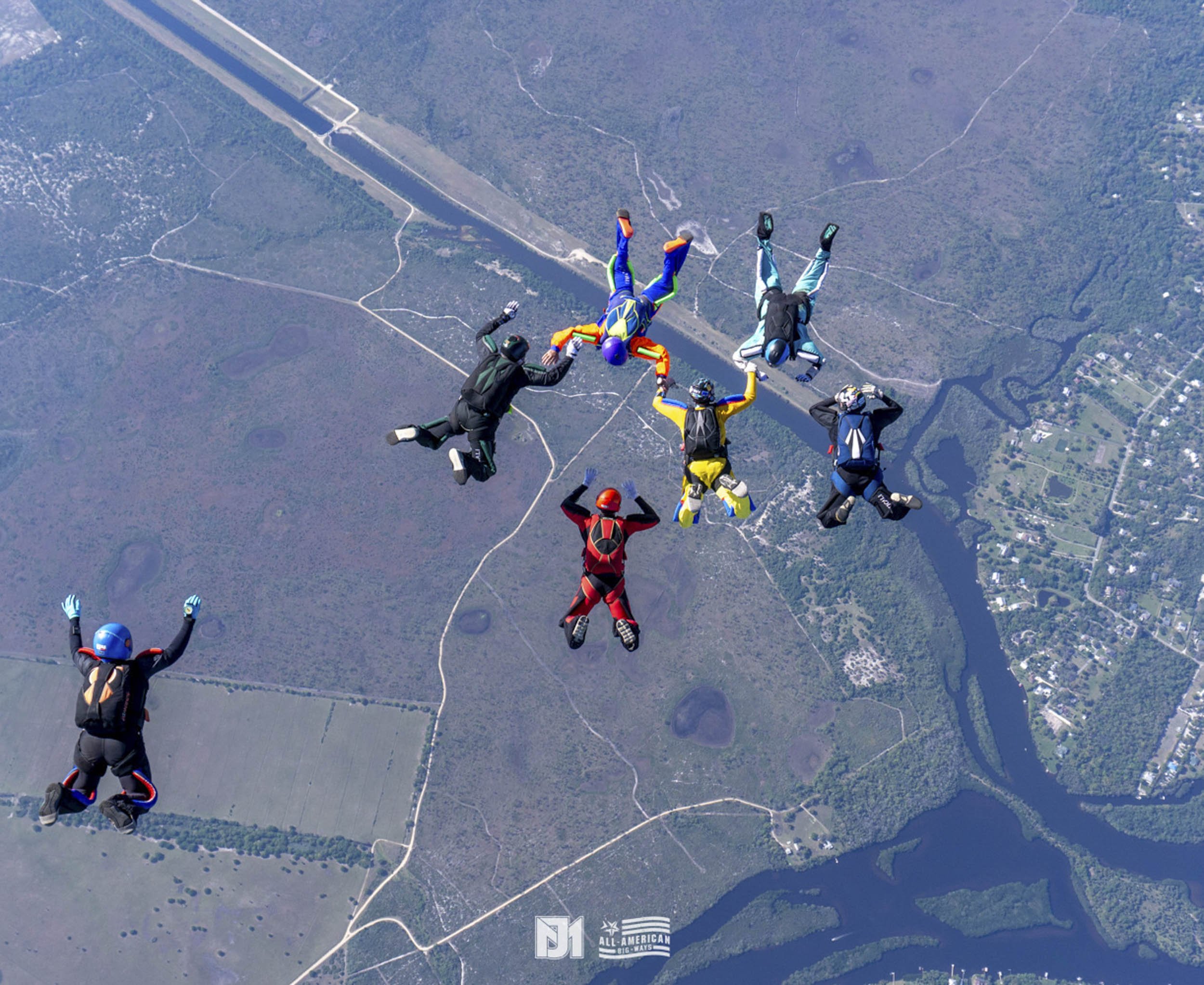Group of skydivers in colorful jumpsuits and helmets forming a star shape during a free fall over a landscape with roads, rivers, and fields.