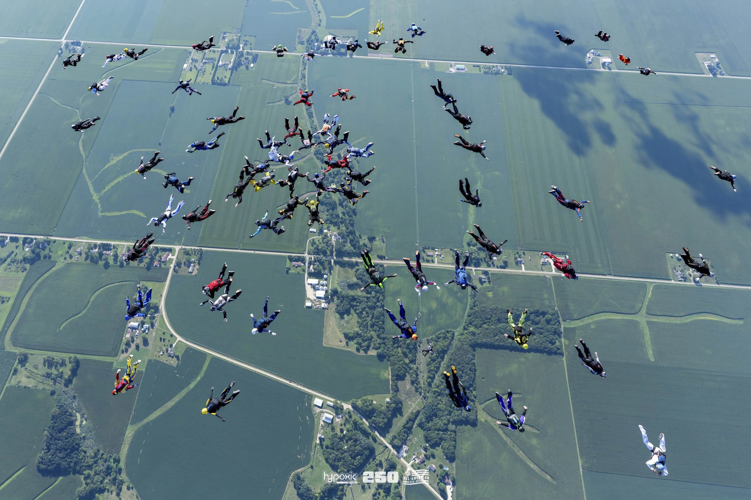 A group of skydivers in colorful jumpsuits and helmets descending with parachutes over farmland and fields.