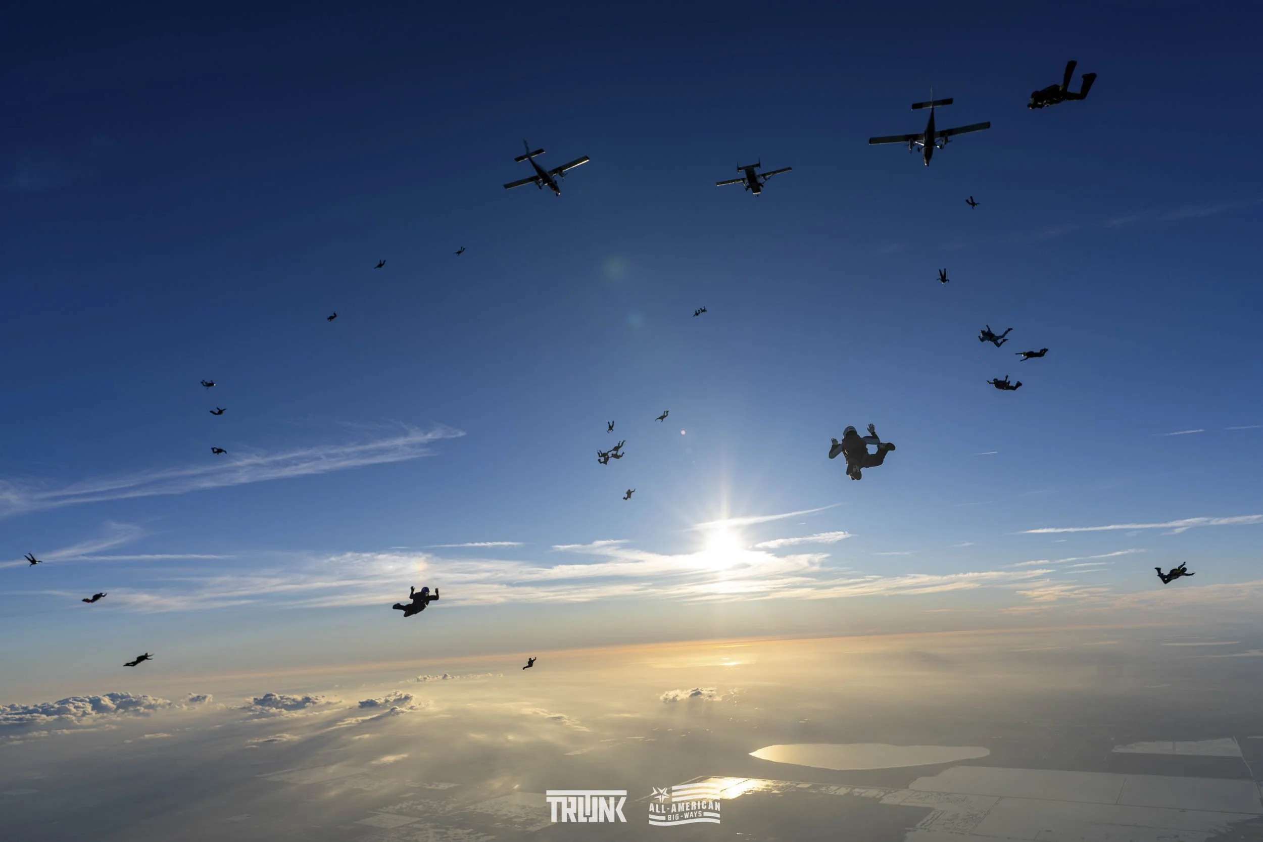 Skydivers free-falling from airplanes at high altitude during daytime with the sun low on the horizon, clouds below, and a clear blue sky.