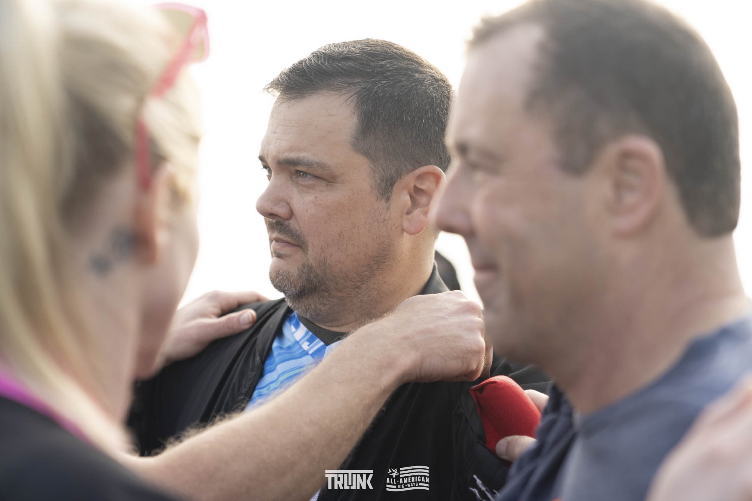 A man with short dark hair, facial stubble, and a serious expression is receiving a supportive gesture from two other individuals, one woman and one man, during an outdoor event. The woman is on the left, partially visible with blonde hair and glasse