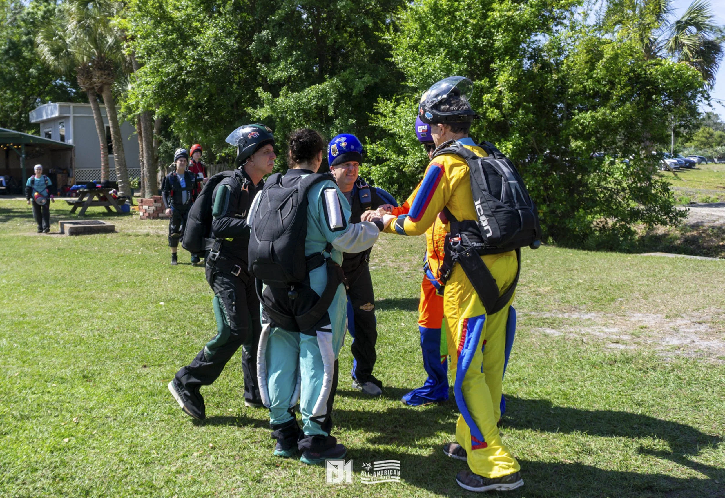 A group of skydivers in jumpsuits and helmets, standing on a grassy field, with some talking and holding hands, with trees and cars in the background.