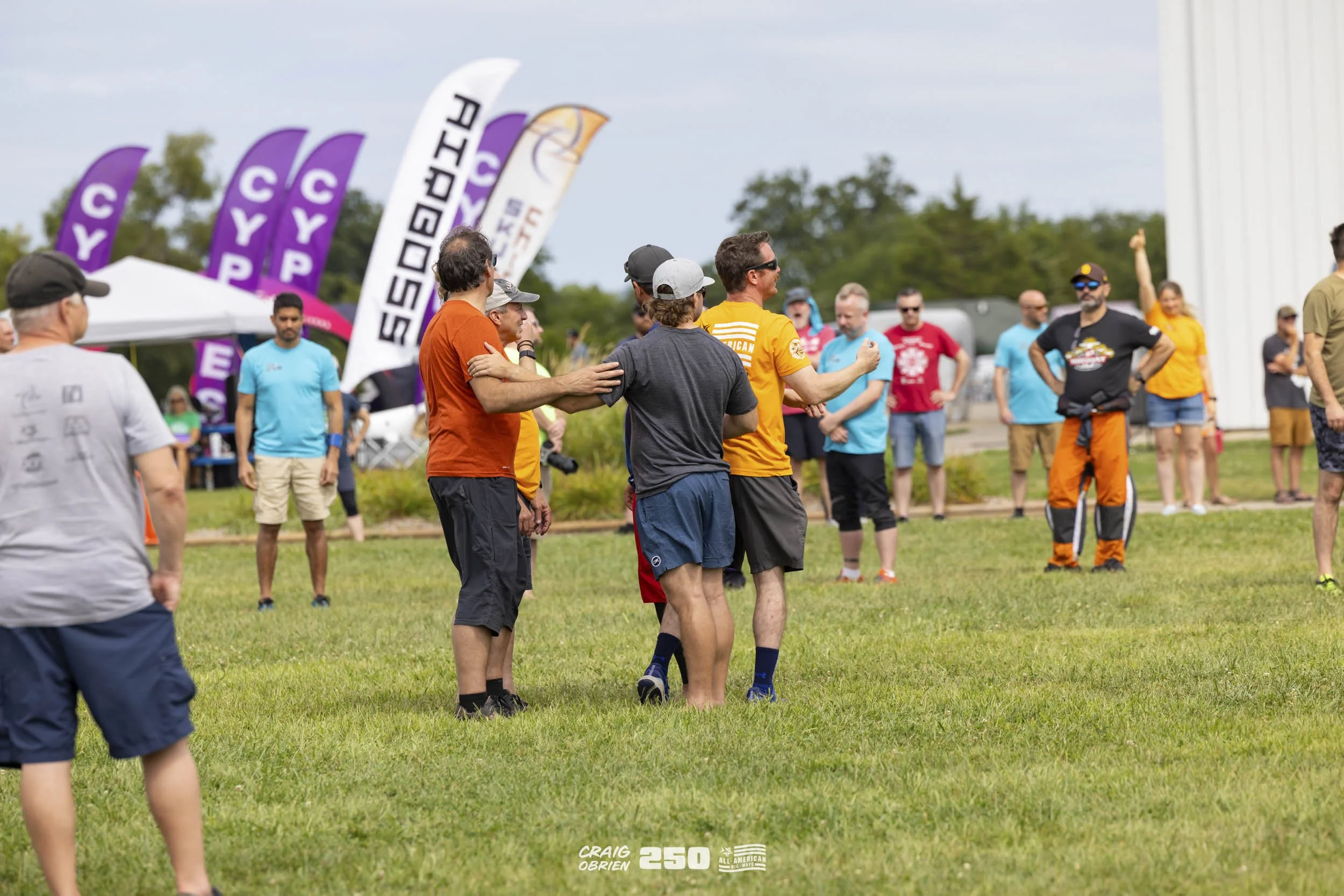 Group of people gathered outdoors on a grassy field, some engaging in conversation, others observing, with flags and tents in the background, during a sporting or community event on a partly cloudy day.