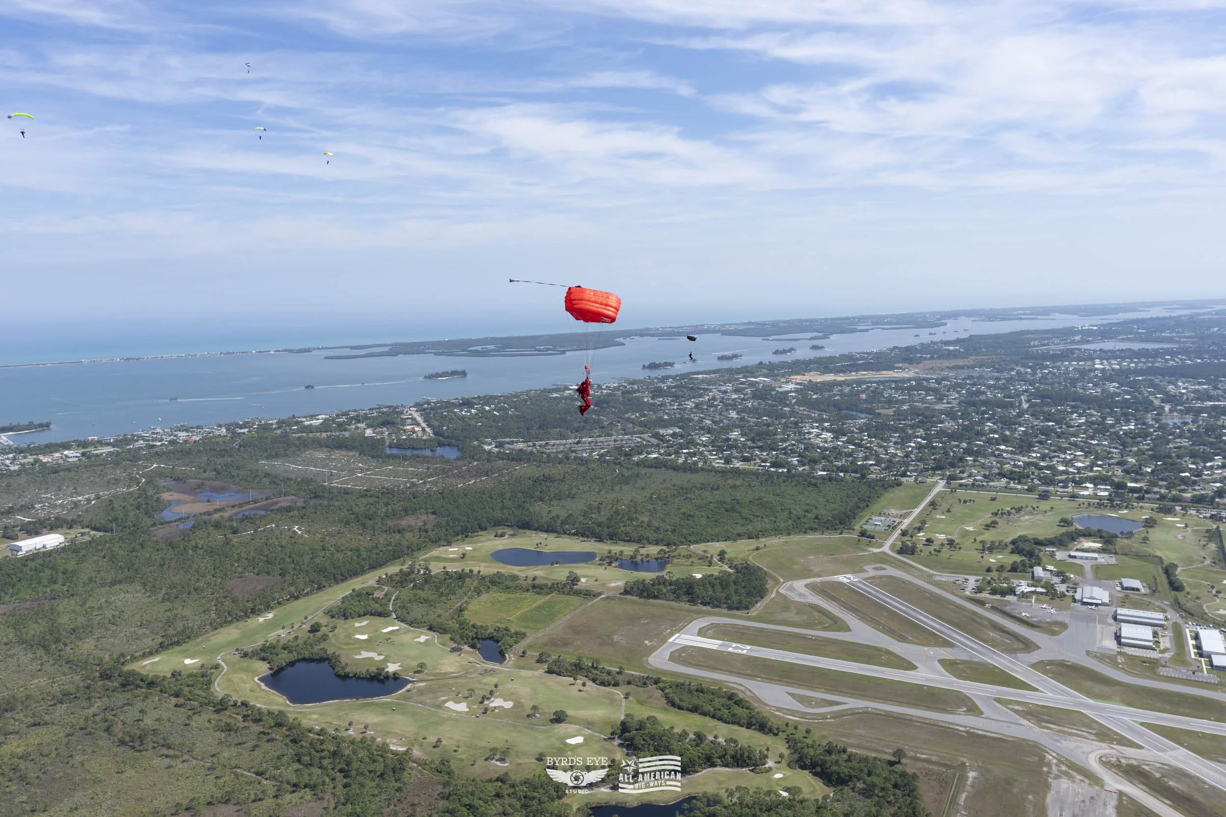 A person in a red jumpsuit with a parachute descending over a landscape with an airport, lakes, and a city in the distance.