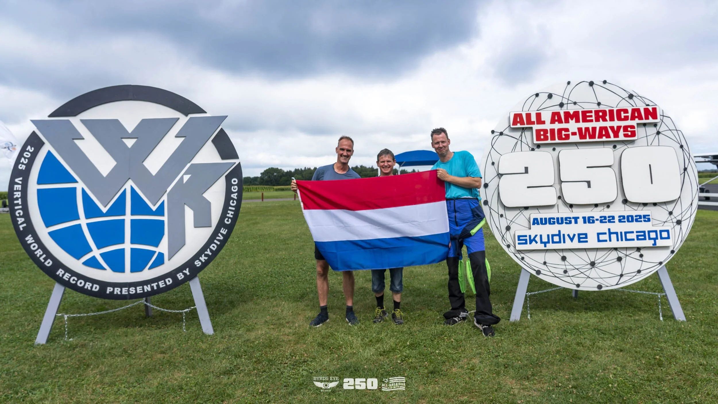 Three people holding a Dutch flag and smiling at the camera, standing between two large signs announcing a world record for skydiving at Skydive Chicago from August 16-22, 2025.