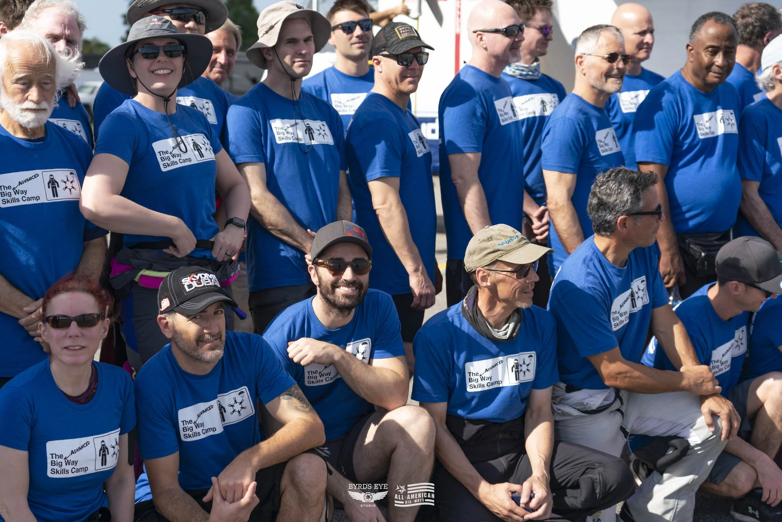 Group photo of people wearing matching blue t-shirts with a logo and text that says 'The Advanced Big Way Skills Camp,' posing outdoors on a sunny day.