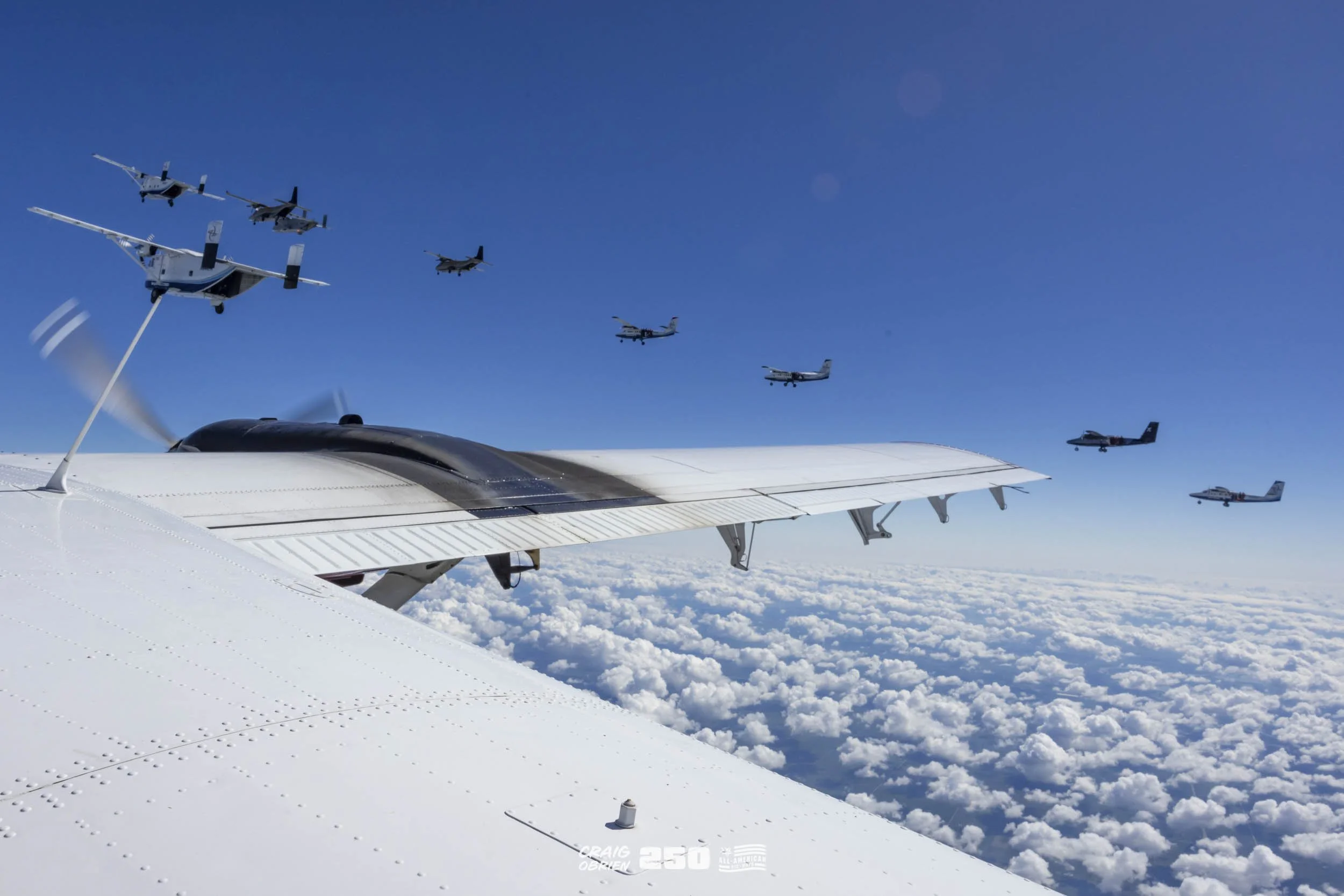 Multiple aircraft flying in formation above the clouds, viewed from the cockpit of a flying airplane.
