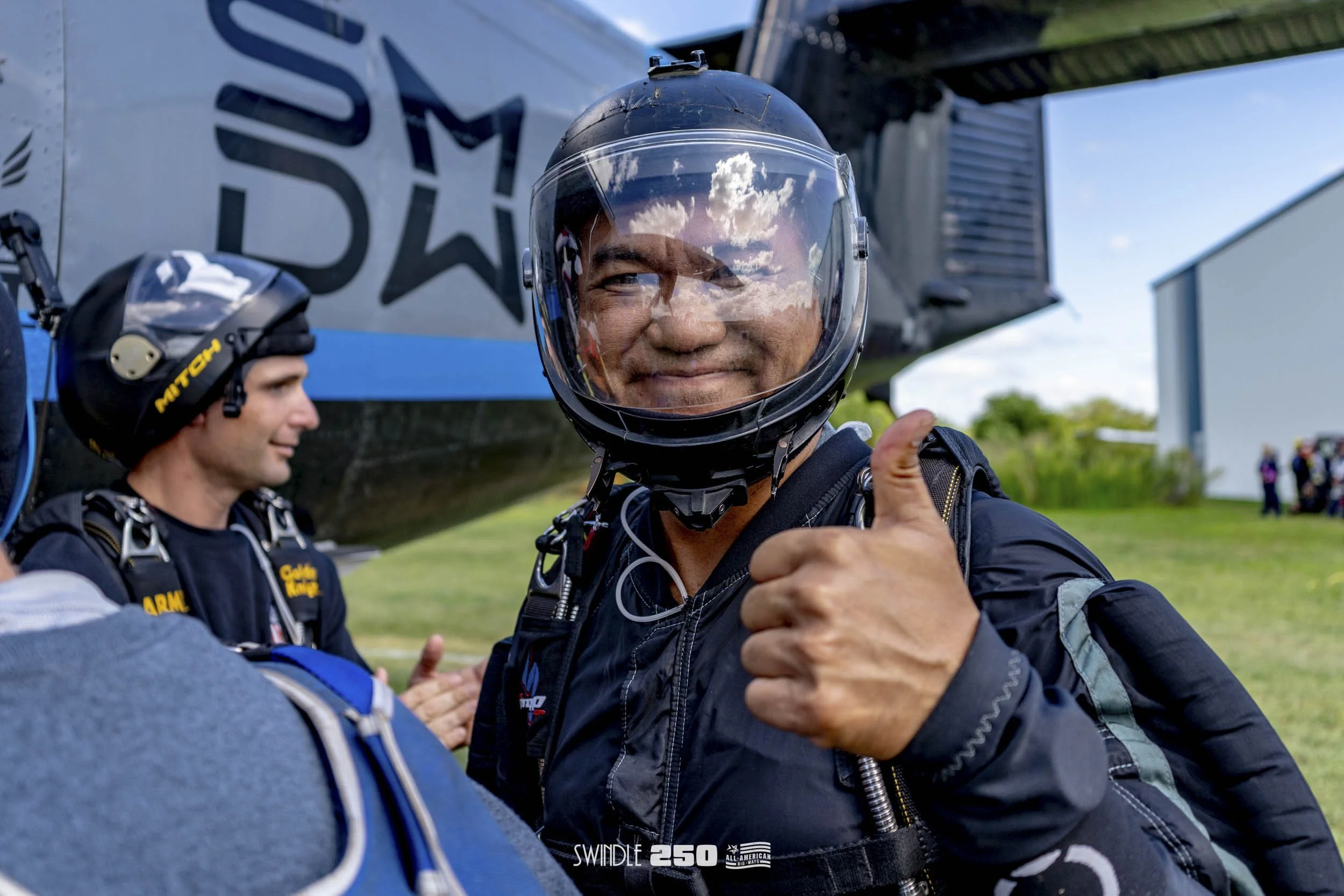 An individual in a helmet and jumpsuit giving a thumbs-up in front of a helicopter, with another person and a group in the background.