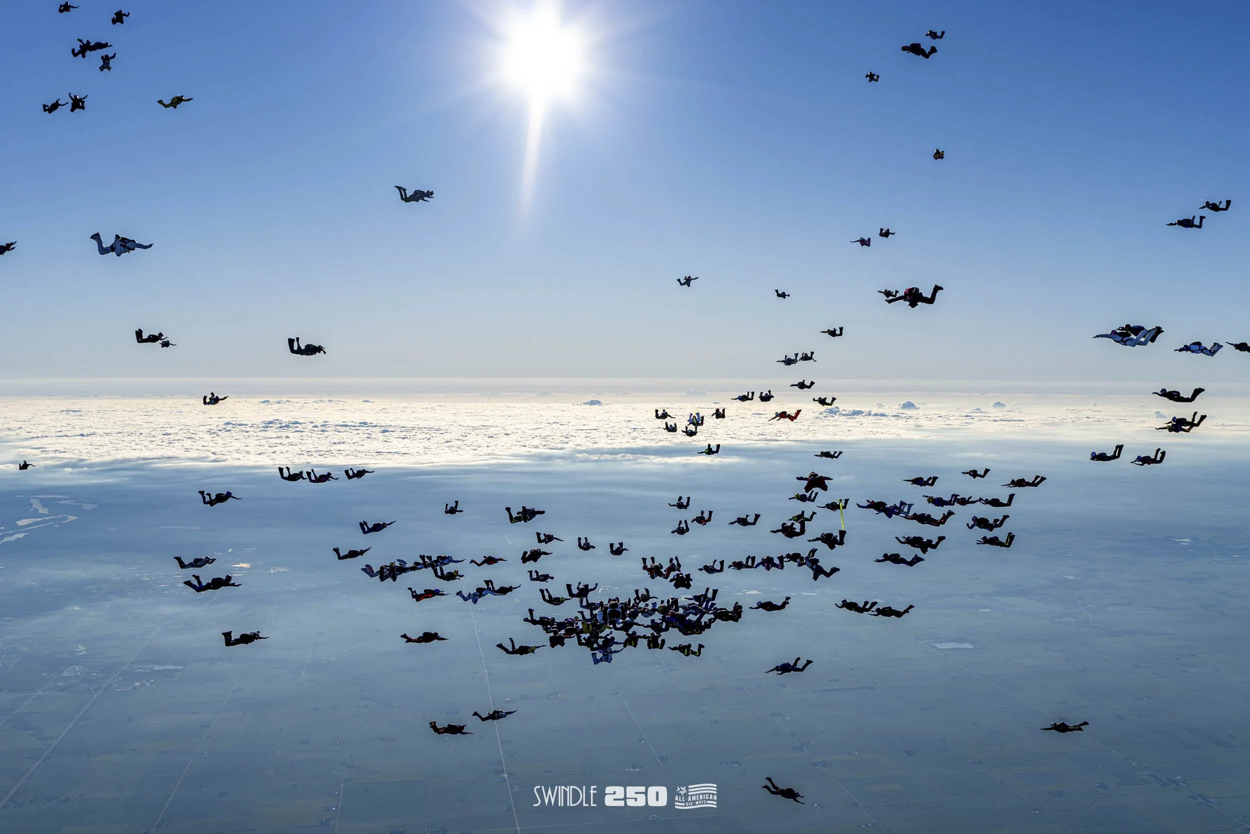 Skydivers jumping in formation over a snowy landscape on a clear day, with the sun shining brightly in the sky.