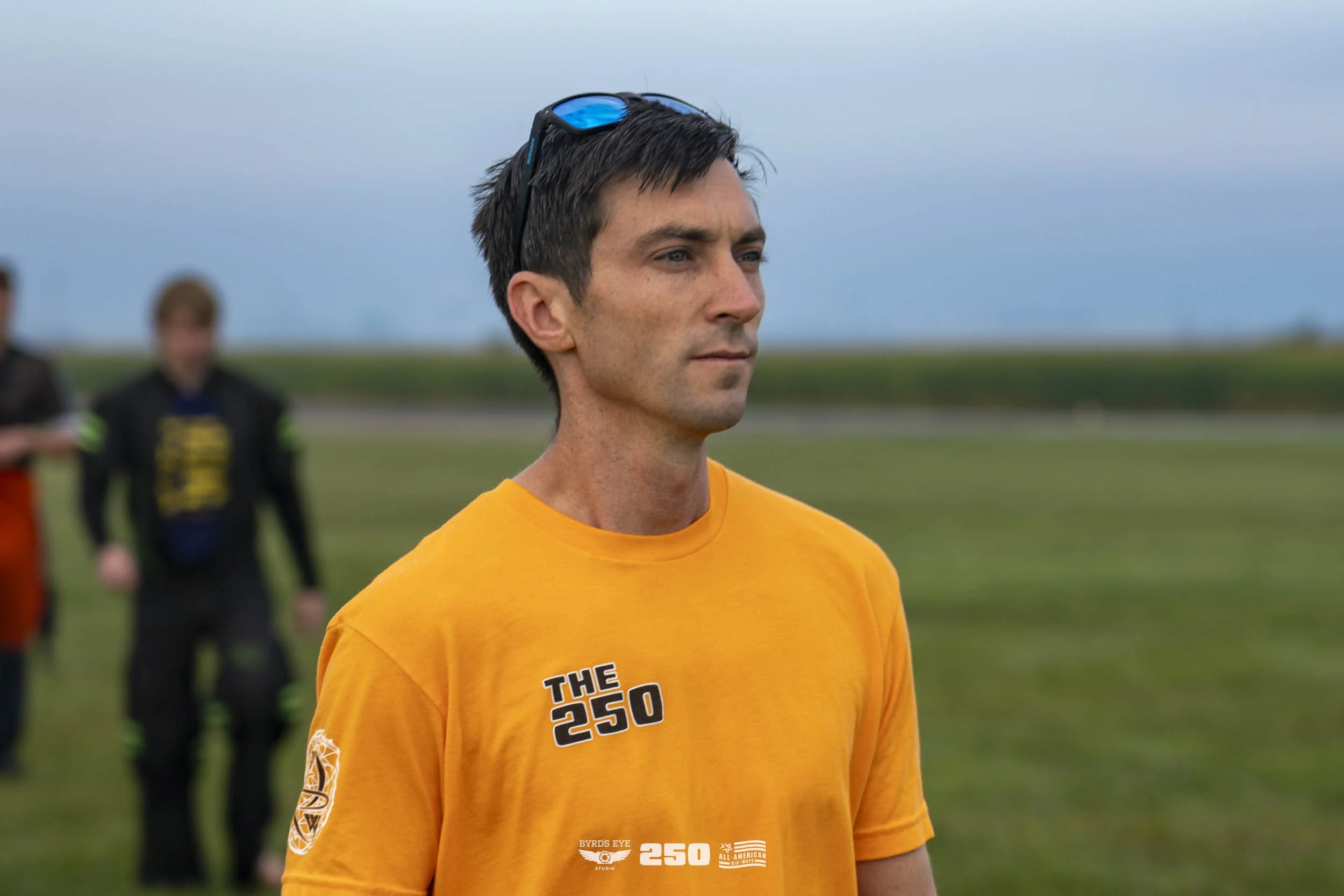 A man with dark hair and blue sunglasses on his head wearing an orange t-shirt with the text 'THE 250' and logos on the front, standing outdoors on a grassy field with a cloudy sky in the background.
