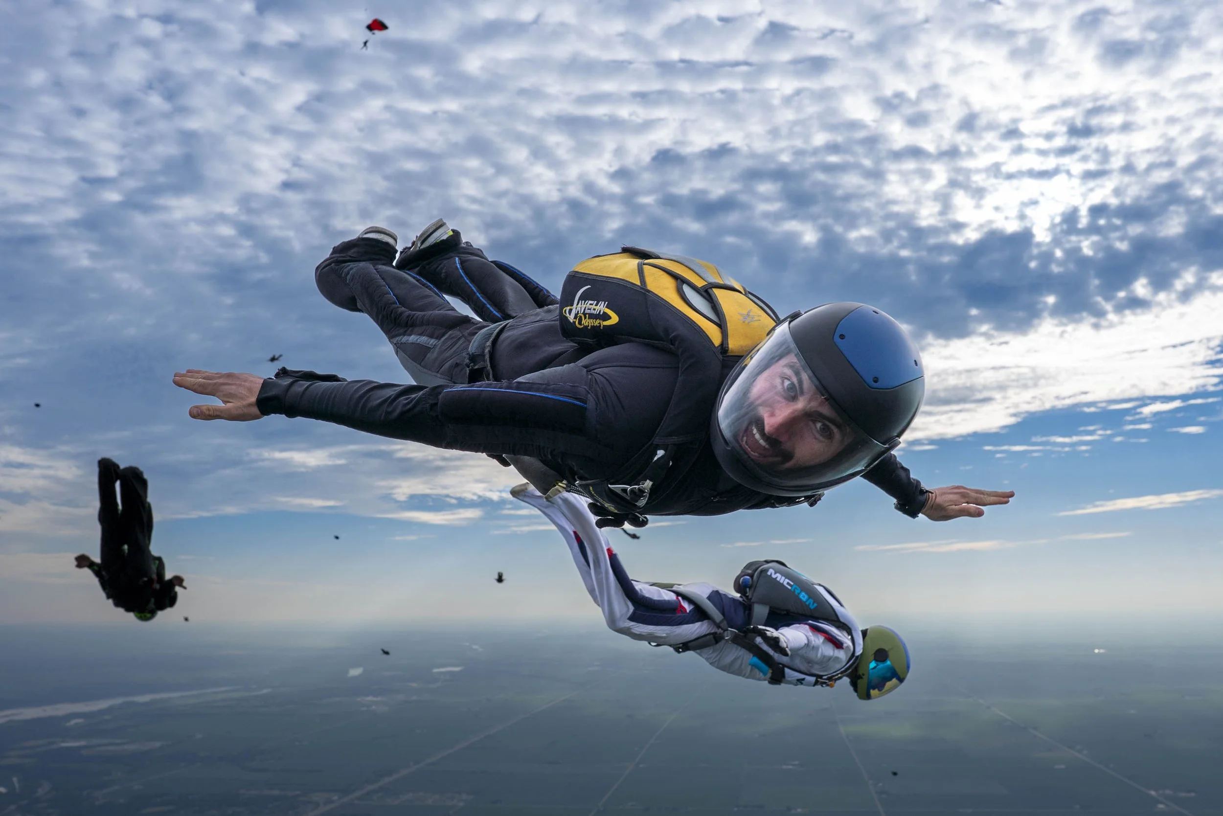 Two skydivers free-falling through the sky with clouds and a landscape below, wearing helmets and jumpsuits, with a yellow backpack on one and a white and blue jumpsuit on the other.