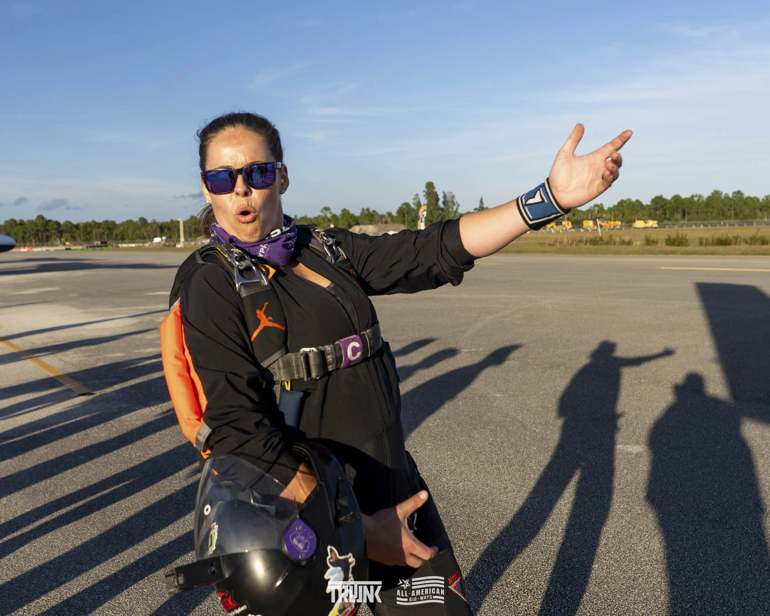 A woman in skydiving gear making a peace sign with her right hand, holding a helmet in her left hand, on an airport tarmac with shadows and a clear sky in the background.
