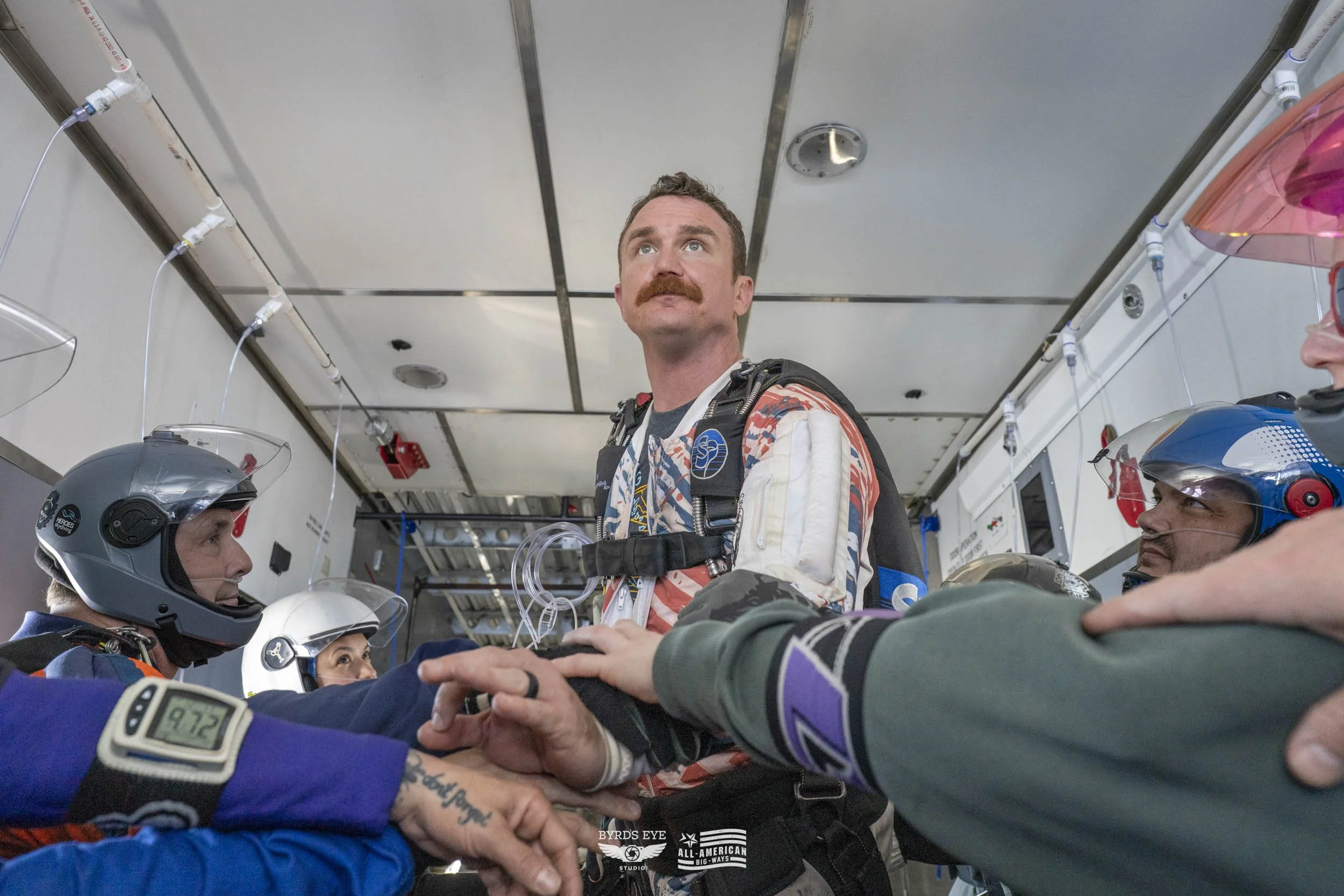 Racing pilots with helmets and gear in a tense pre-race moment inside the aircraft or pit crew area, focused on a central driver standing with team members surrounding him.