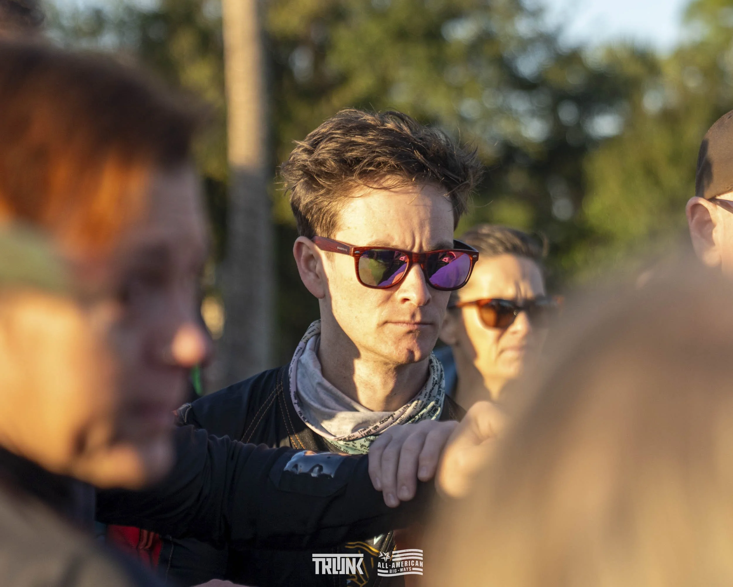 Group of people outdoors, some wearing sunglasses, with trees in the background, sunlight, serious expressions, focus on a young man with brown hair and purple-tinted sunglasses.