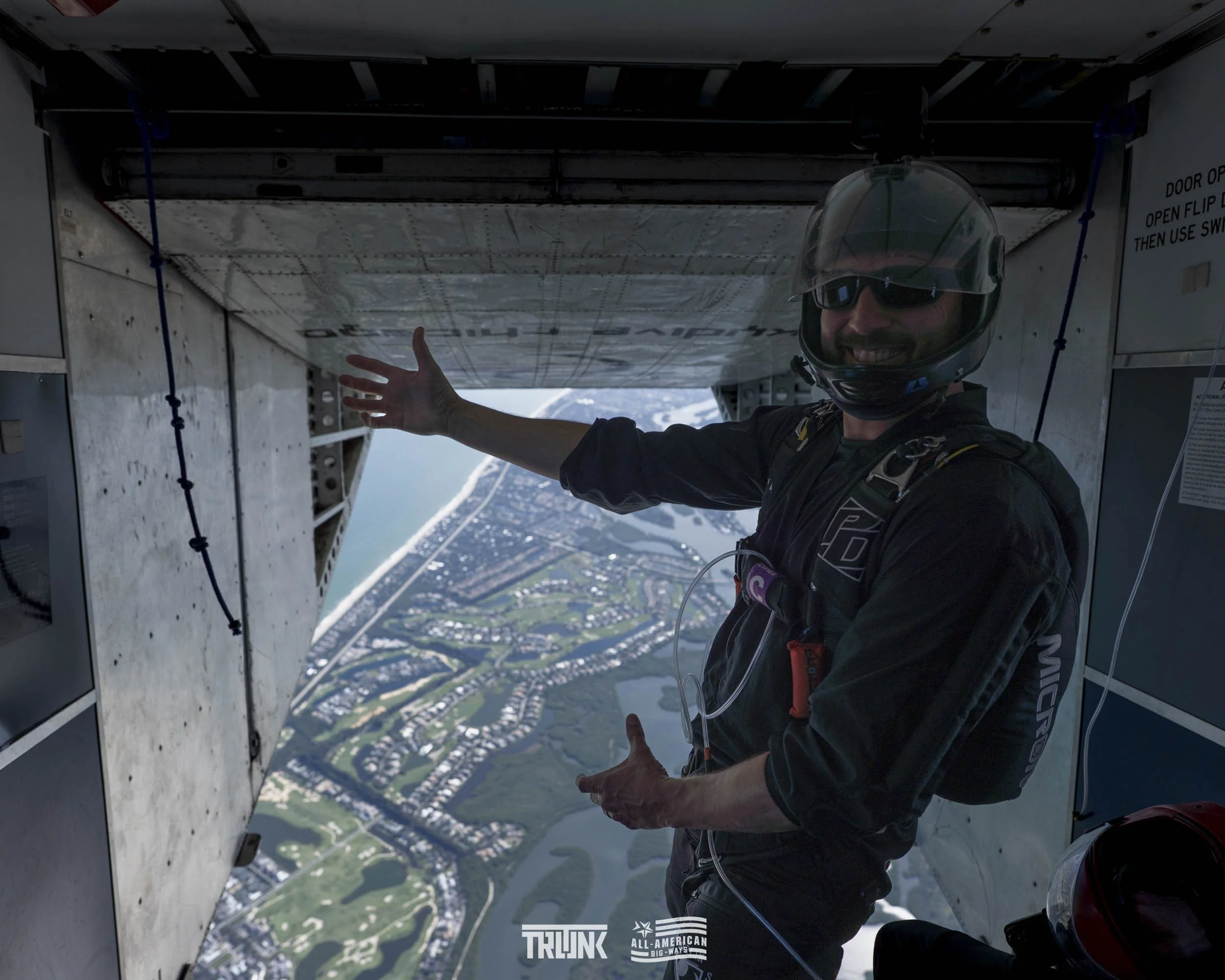 A skydiver inside an aircraft, wearing a helmet and sunglasses, pointing outside with a bright smile. The open aircraft door reveals a view of a coastal landscape with water, green fields, and residential areas.