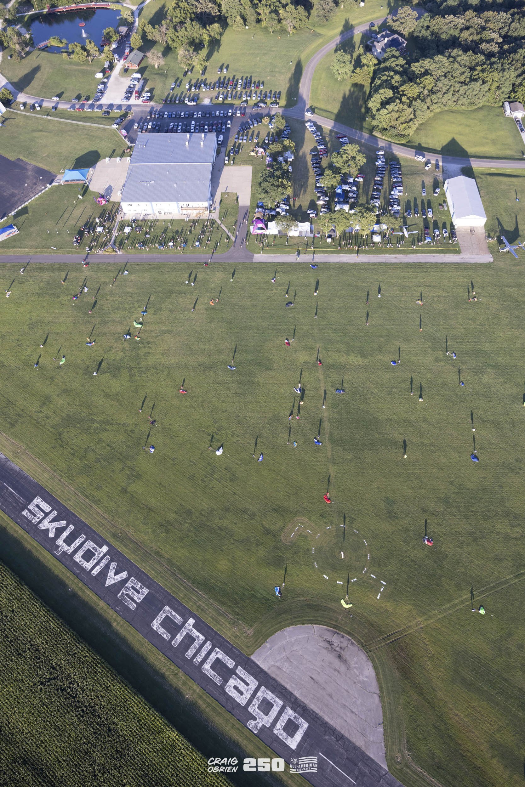 An aerial view of a large outdoor event featuring numerous parachutists landing on a grassy field marked with 'DODGE CHALLENGE' in large letters, with a building, parking area, and trees in the background.