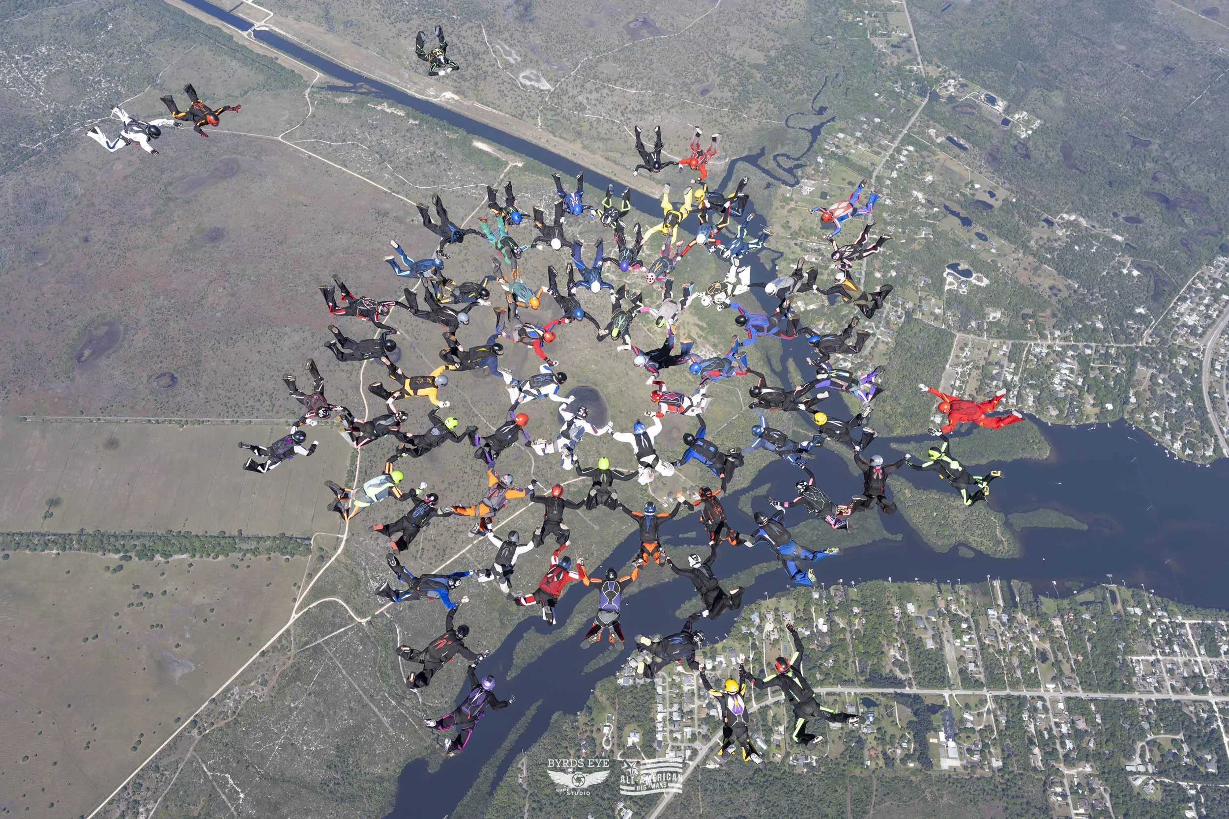 Skydivers in free fall formation over a landscape with lakes and a river.