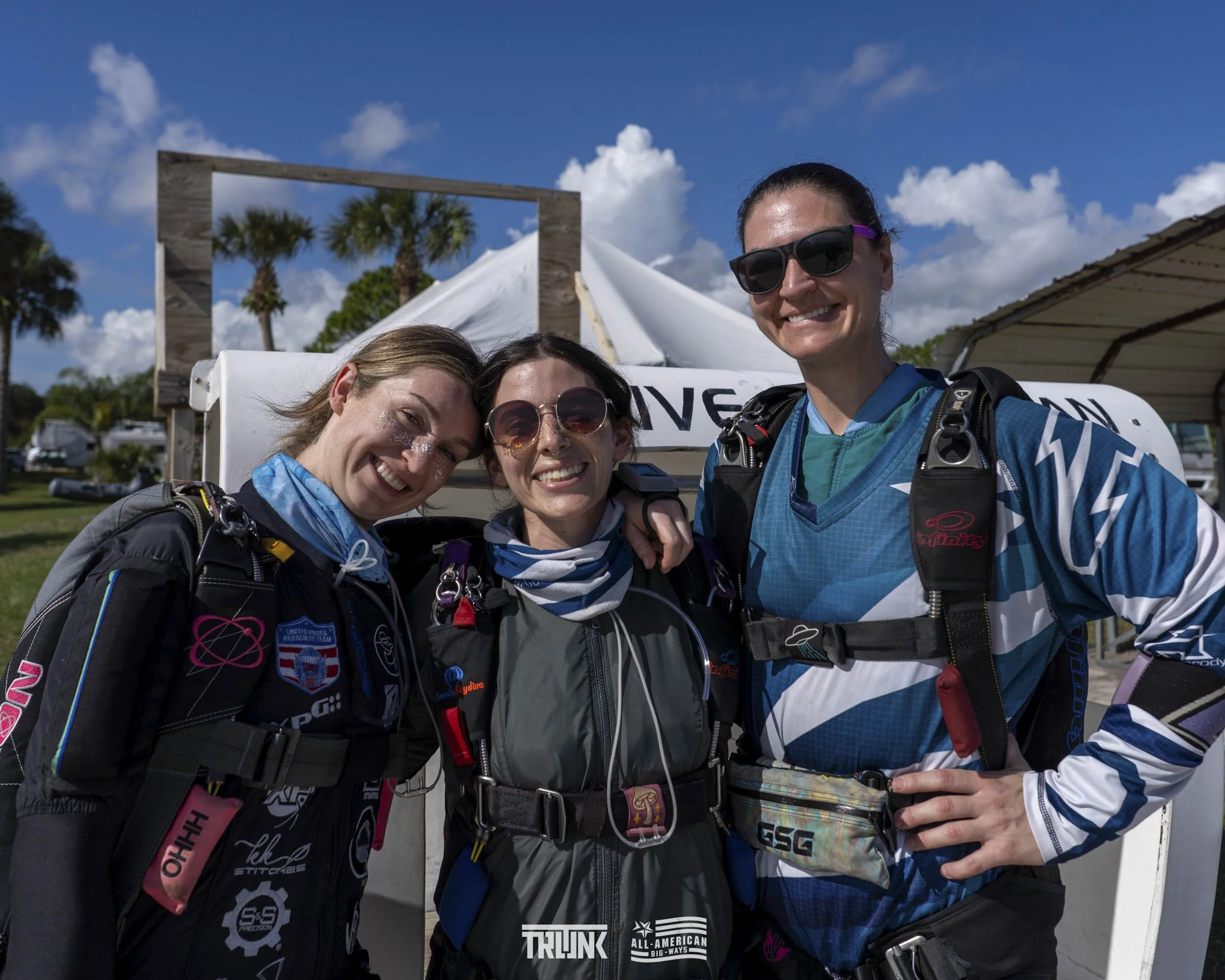 Three women in skydiving jumpsuits smiling, standing close together with their arms around each other, with skydiving gear, in front of a tent and palm trees on a sunny day.