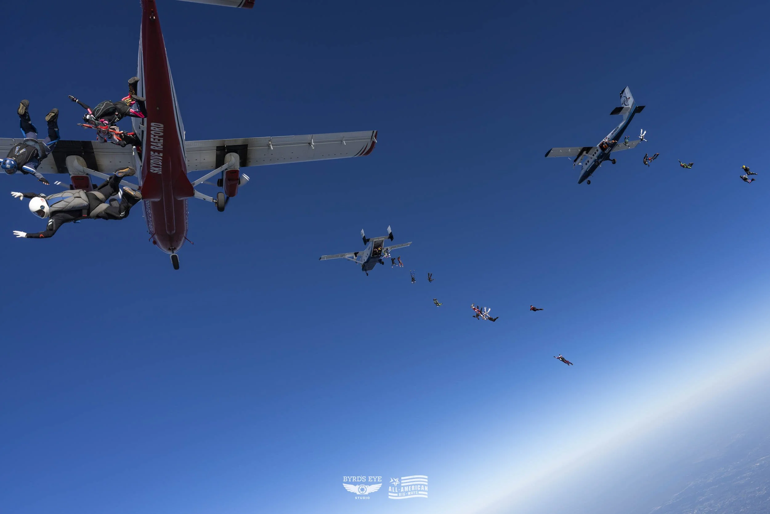 Multiple skydivers jumping from airplanes against a clear blue sky.
