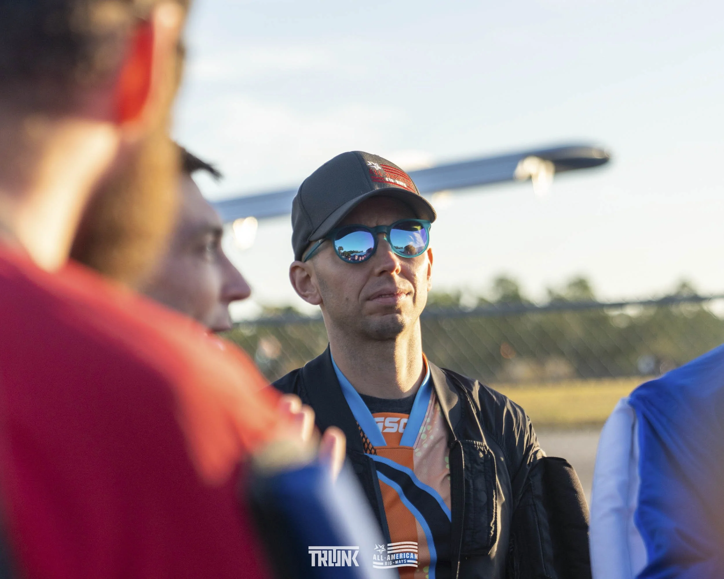 A man wearing mirrored sunglasses, a gray baseball cap, and a racing jacket with a medal around his neck stands with others on a racetrack outdoors during daytime.