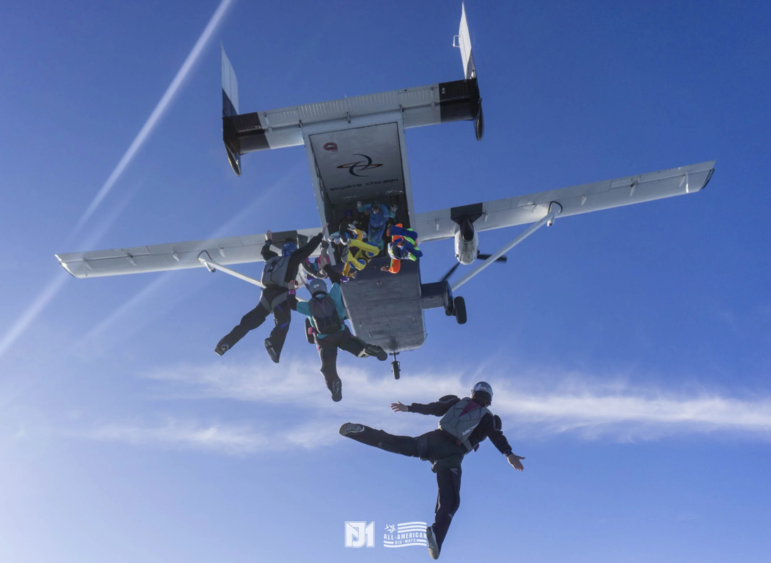 Skydivers exiting a small aircraft in free fall against a blue sky, with some trailing smoke.