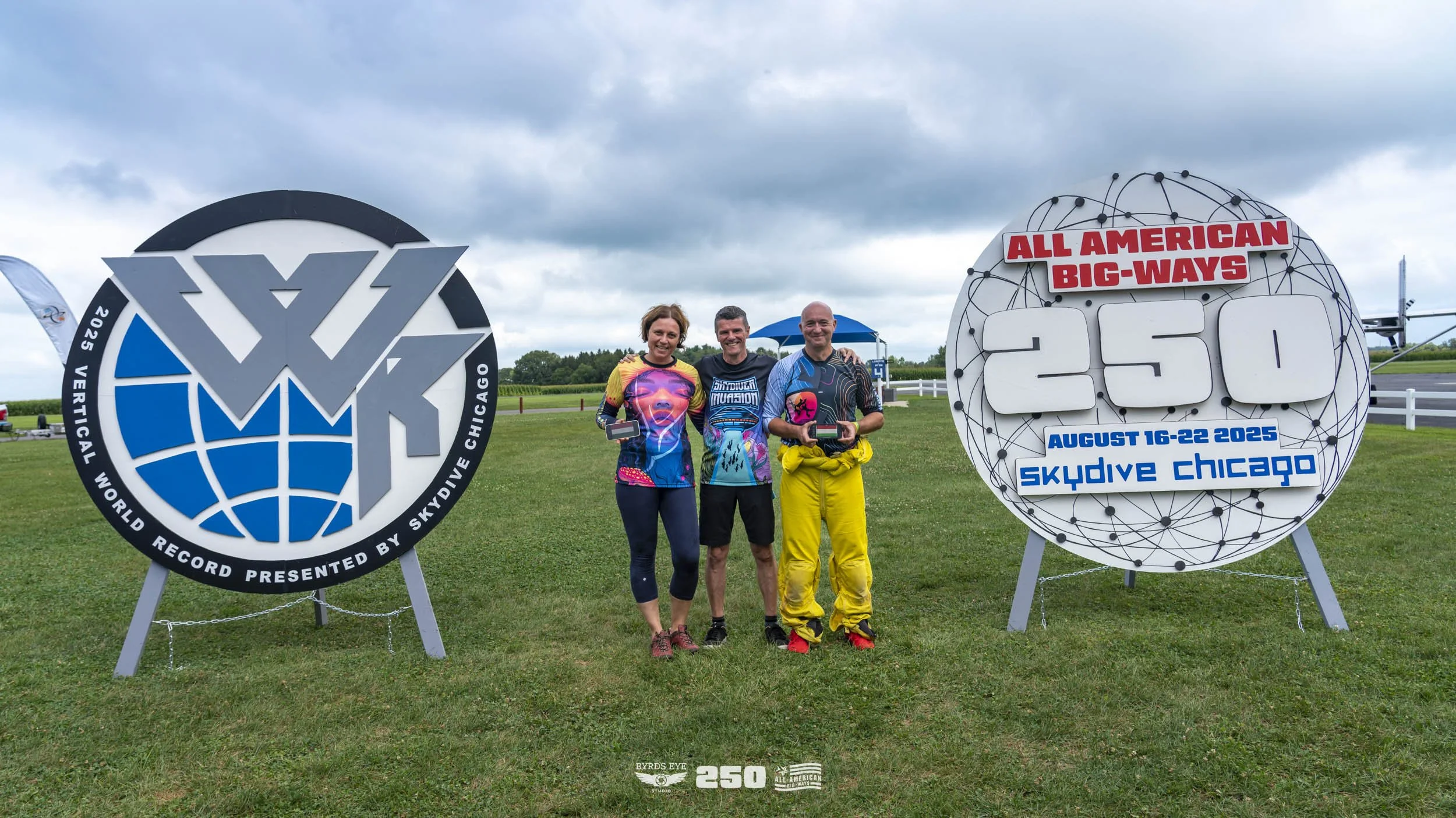 Three people standing on grass, holding awards after a wingsuit record attempt at Skydive Chicago, August 16-22, 2025. Two oversized event signs are behind them, one displaying the wingsuit record and the other showing the event details.