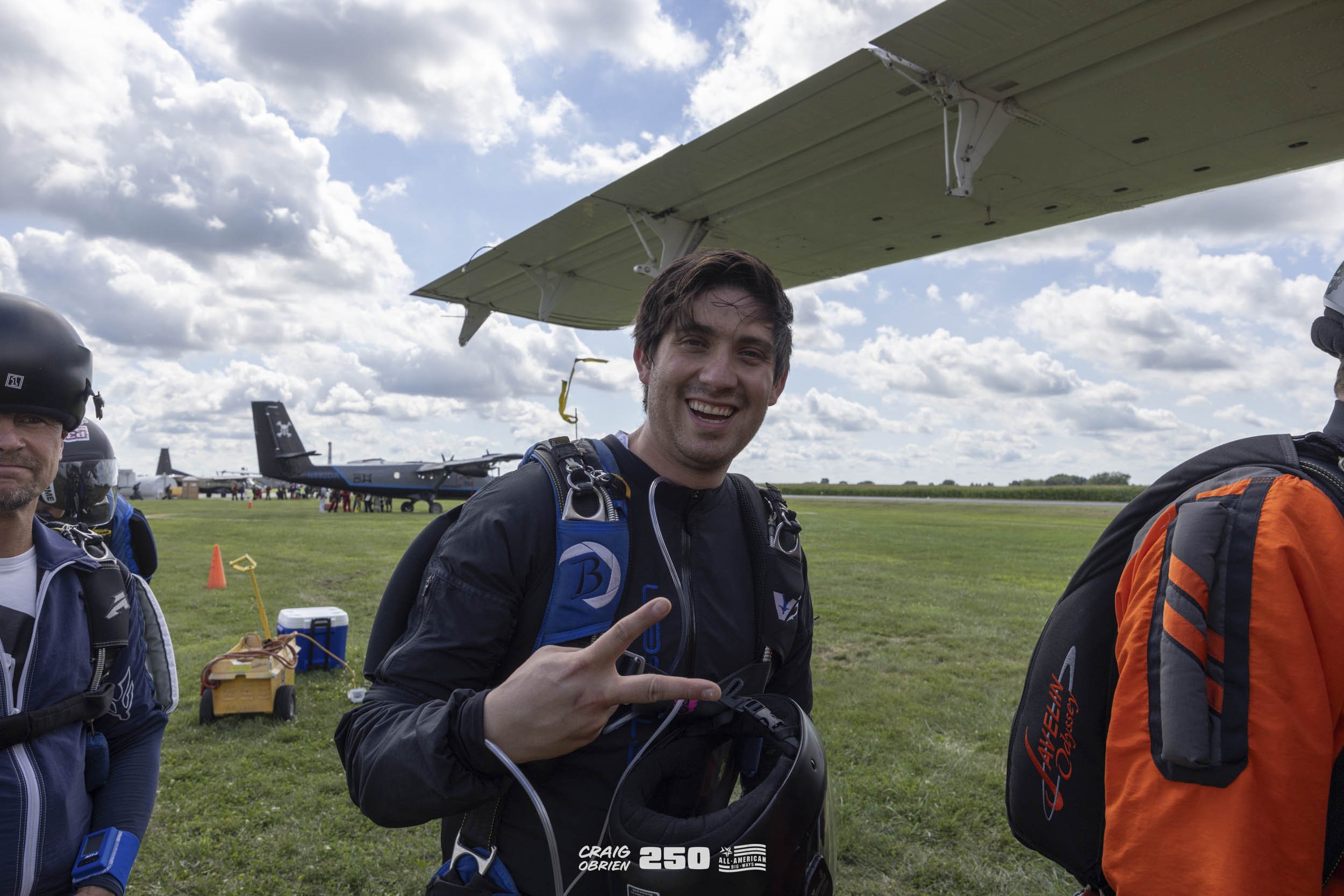 A man in skydiving gear smiling and making a peace sign while standing outdoors near an open field with aircraft and skydiving equipment in the background.