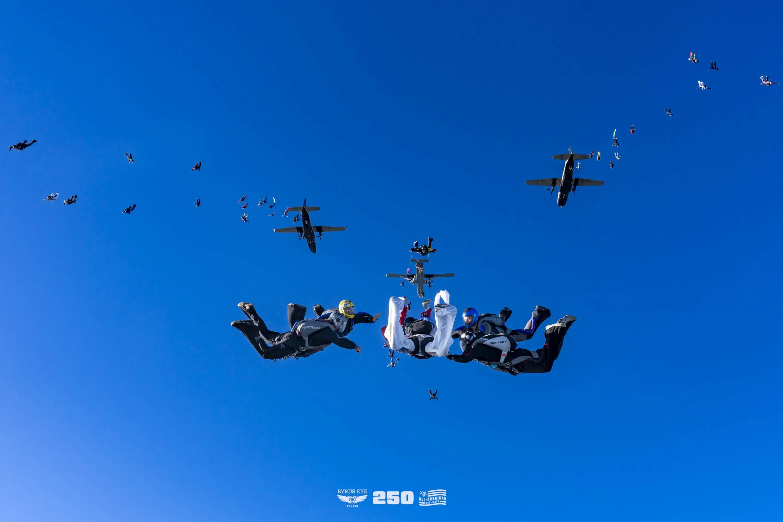 Skydivers in freefall formation with two aircraft flying in the background against a clear blue sky.