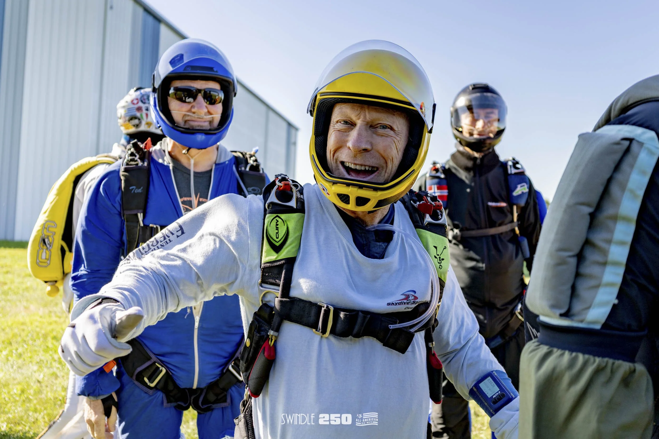 Group of skydivers in jumpsuits and helmets preparing for a jump outdoors on a sunny day.