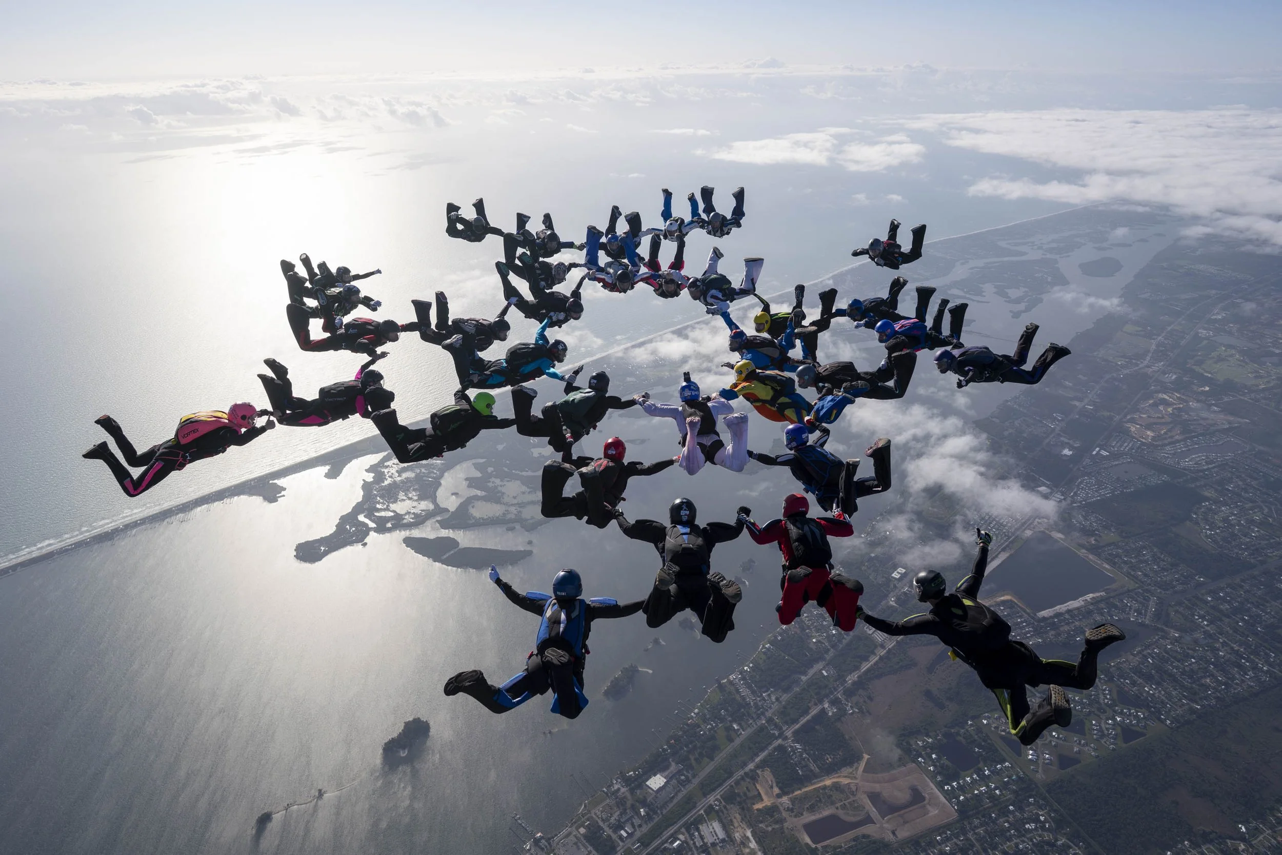 Group of skydivers holding hands in formation mid-air over a body of water and land, with some clouds in the sky.