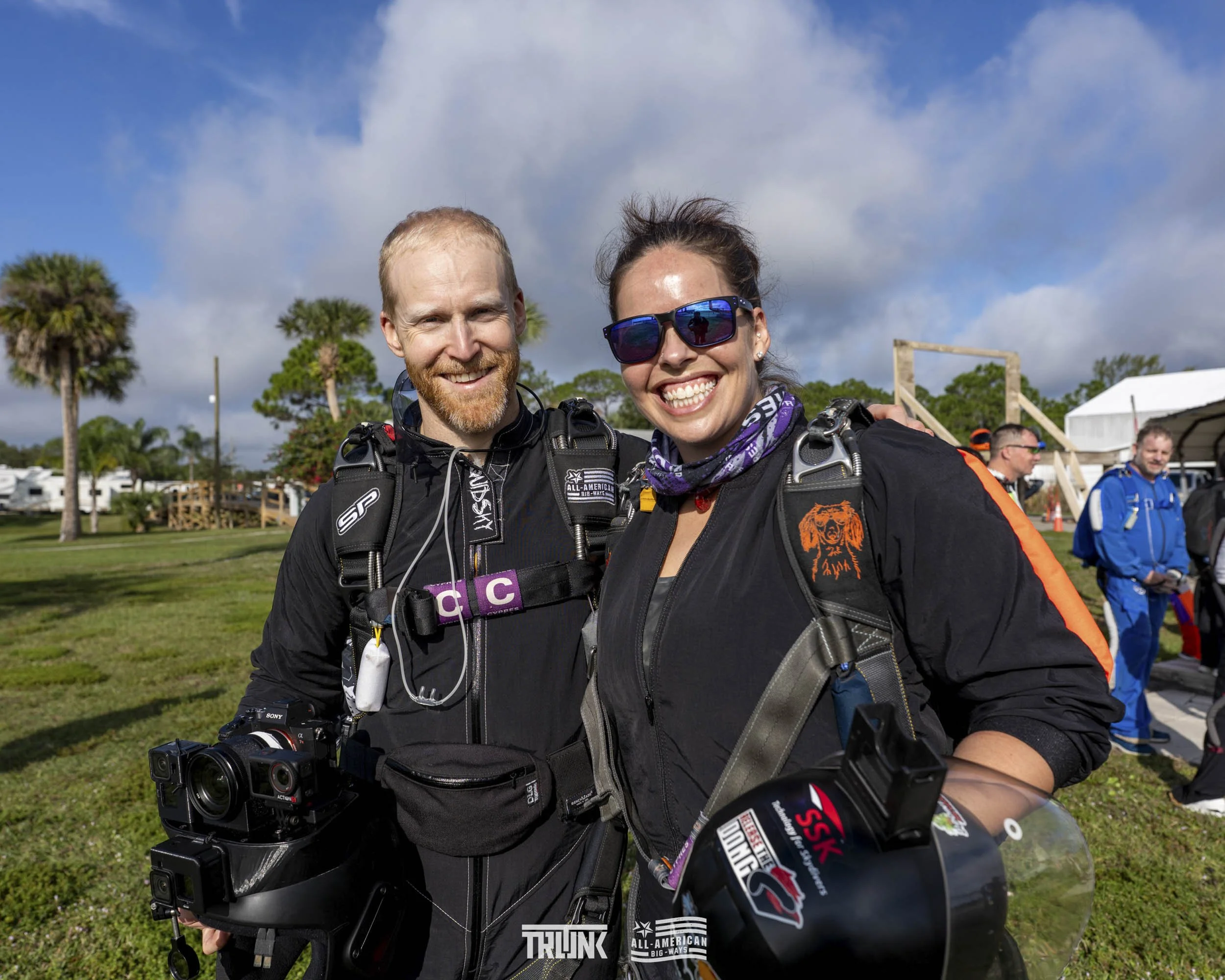 Two smiling skydivers in black jumpsuits, standing outdoors with other skydivers and a white tent in the background, under a partly cloudy sky.