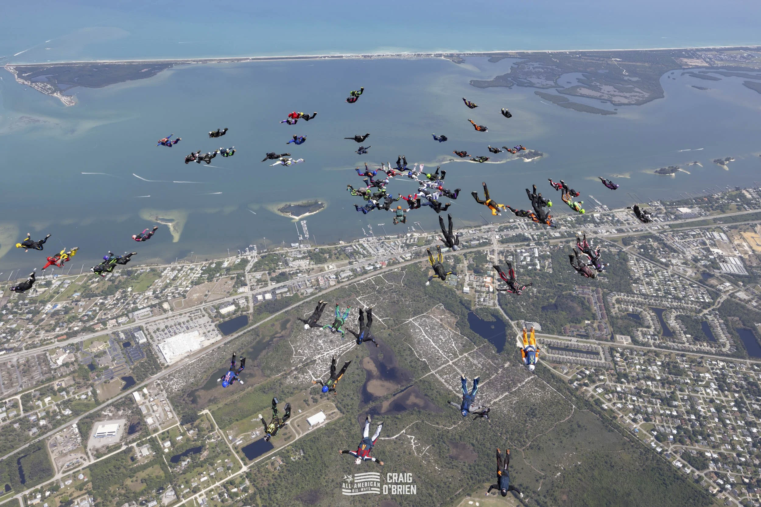 A large group of skydivers in colorful jumpsuits and gear jumping out of an airplane over a landscape with water, islands, and a city.