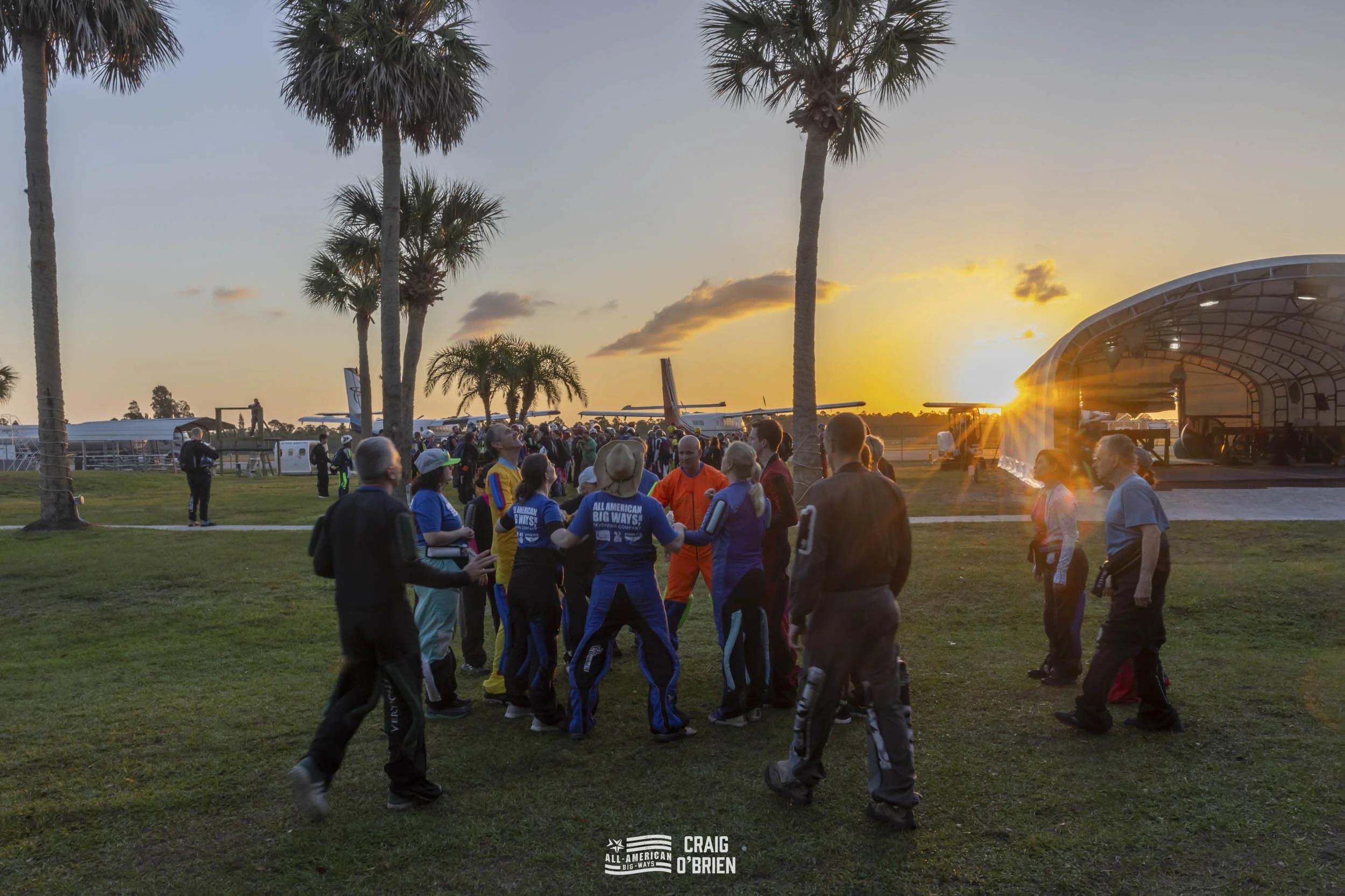 People gathered outdoors near palm trees during sunset at an airport, with airplanes in the background and a stage on the right.