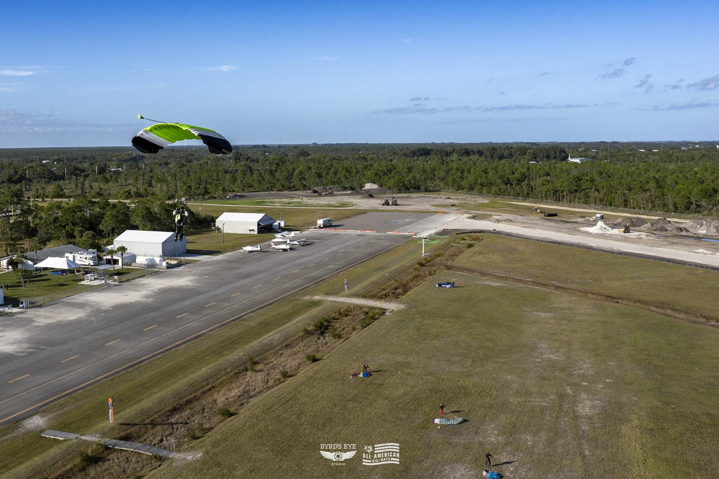 A person is flying in a green and black parachute above a small airport runway with several small aircraft parked nearby, and some people on the ground preparing for a parachute jump on a clear day.