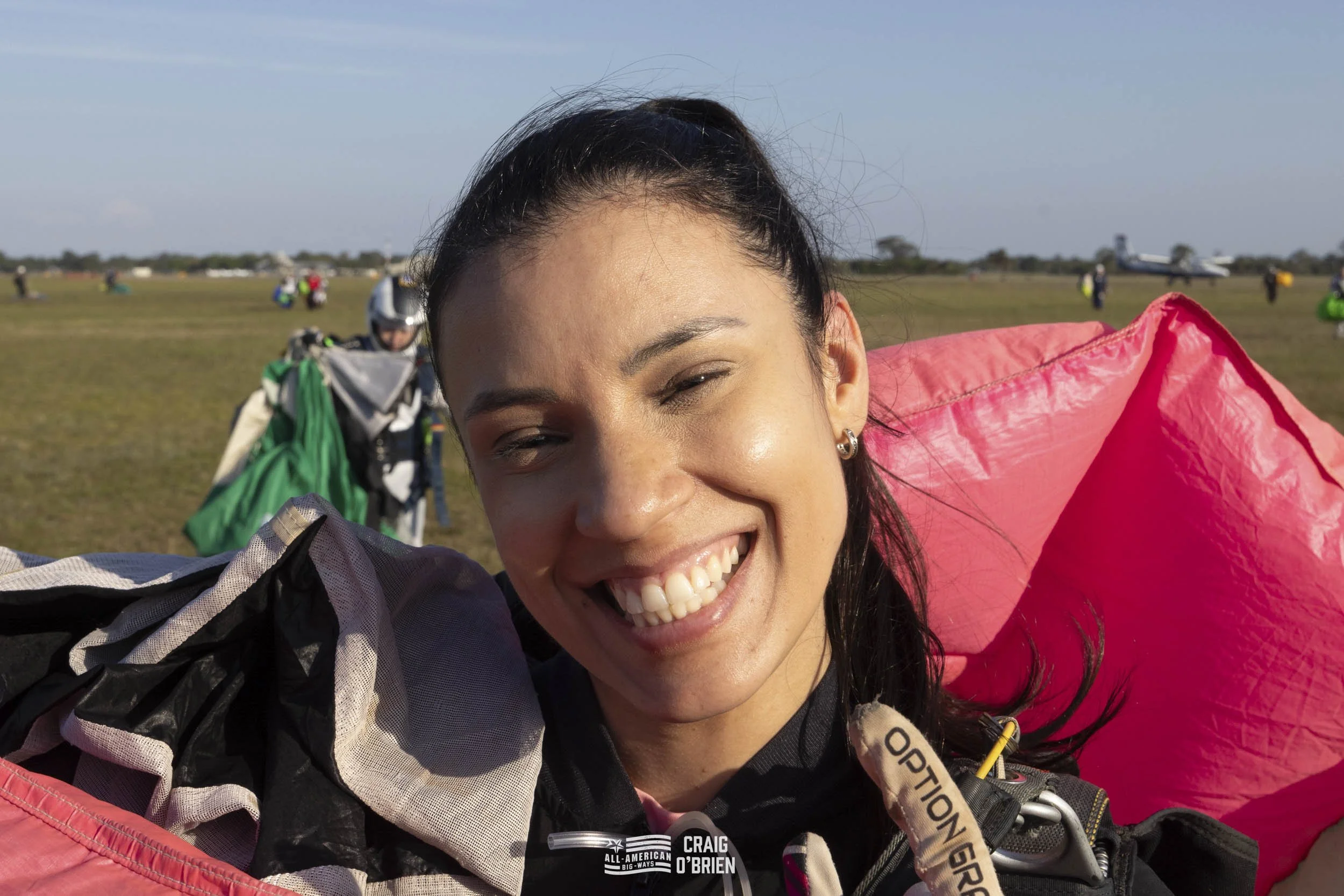 A woman smiling after a skydiving jump, wearing a jumpsuit and gear, with an open field and small airplane in the background.