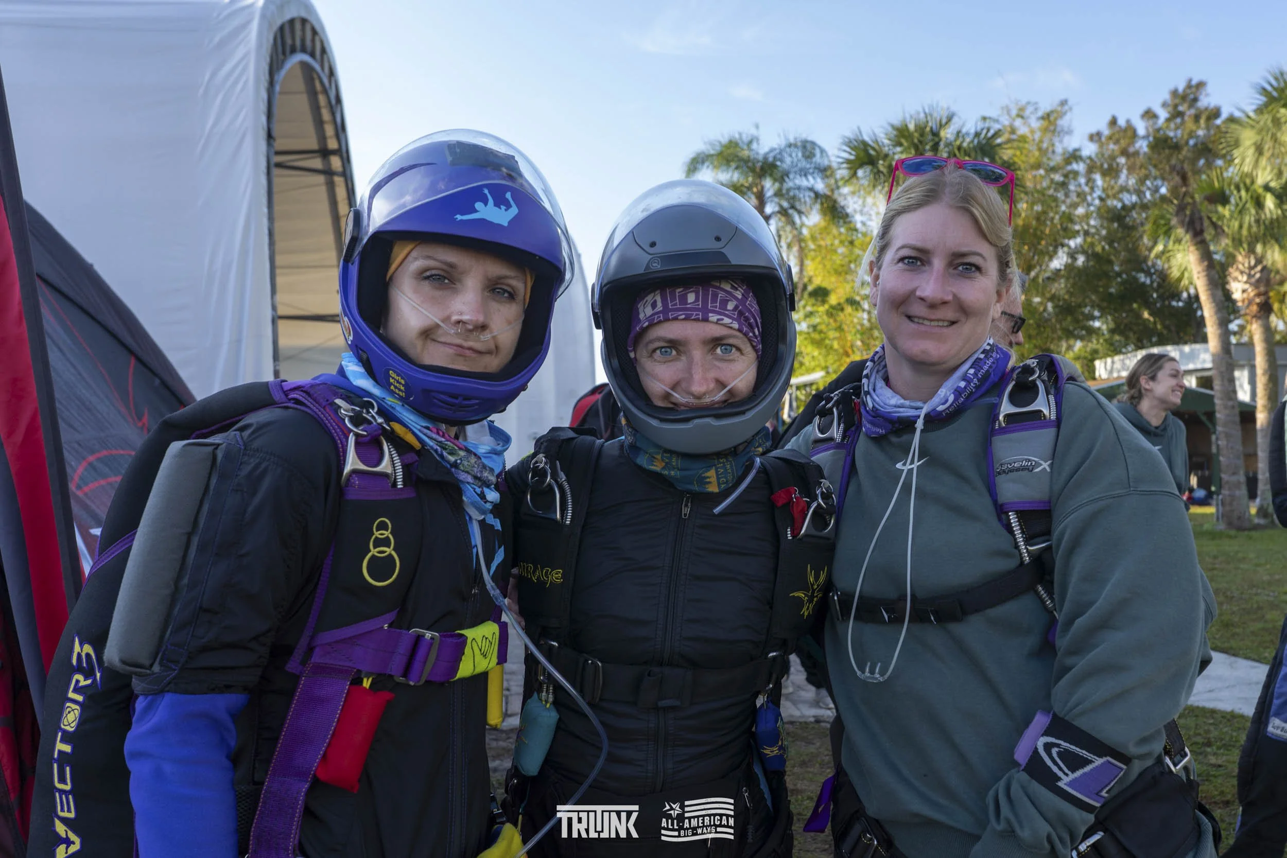 Three women dressed in skydiving gear, wearing helmets and harnesses, standing outdoors in front of trees and a tent, smiling at the camera after a skydiving jump.