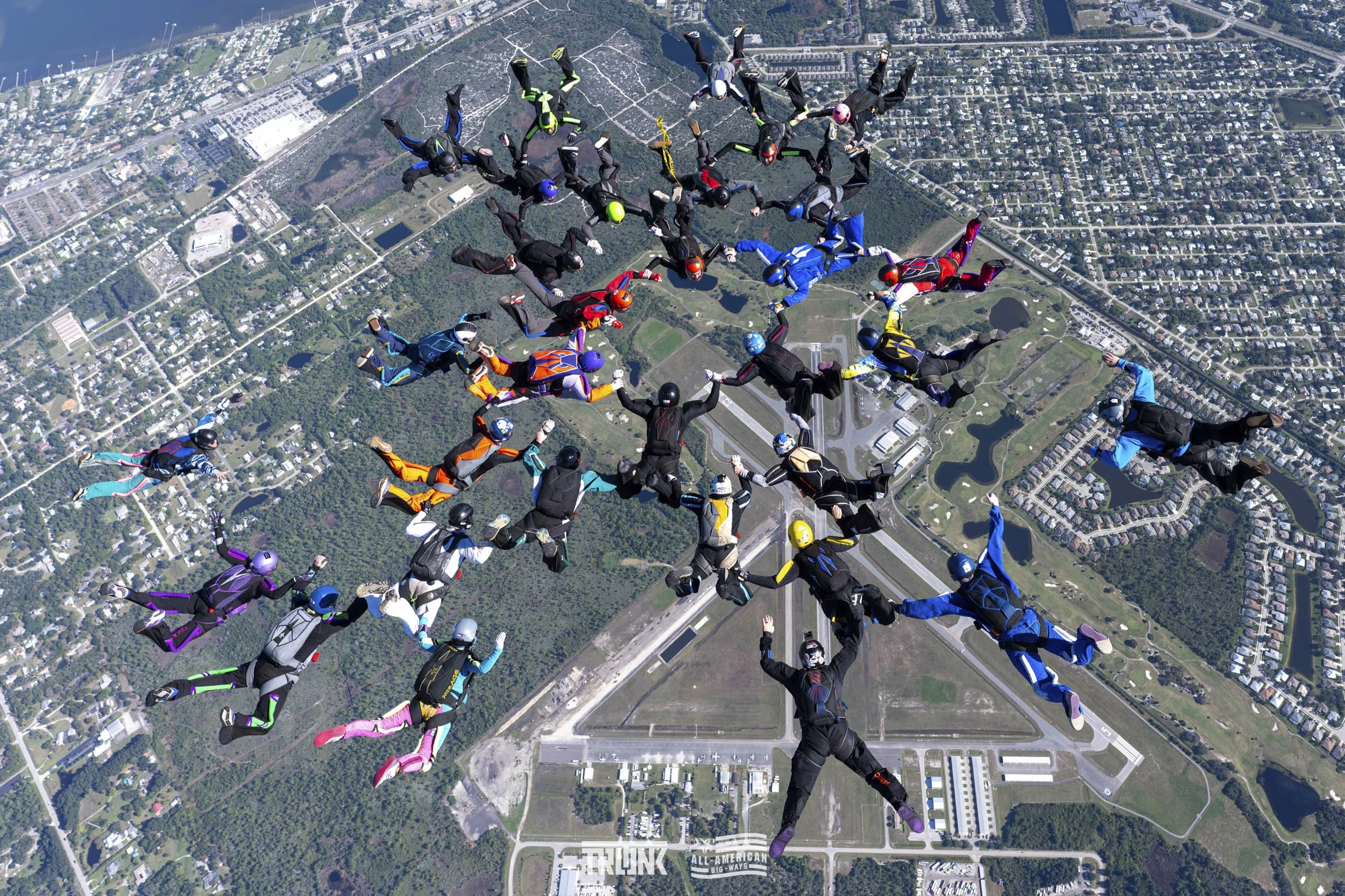 Group of skydivers in colorful jumpsuits and helmets forming a heart shape over a landscape with roads, trees, and buildings.
