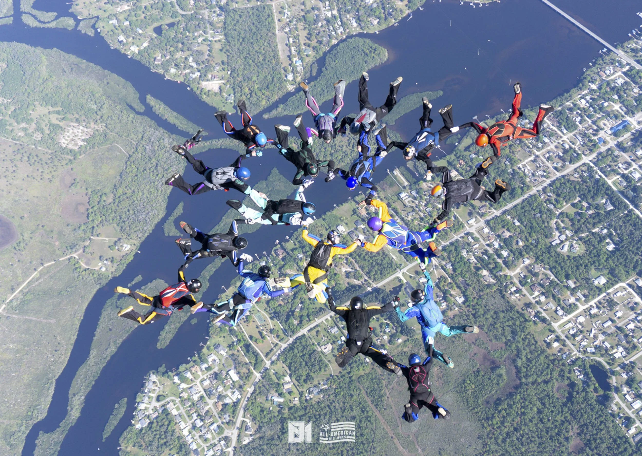 Group of skydivers forming a star shape in freefall over a landscape with lakes and trees.