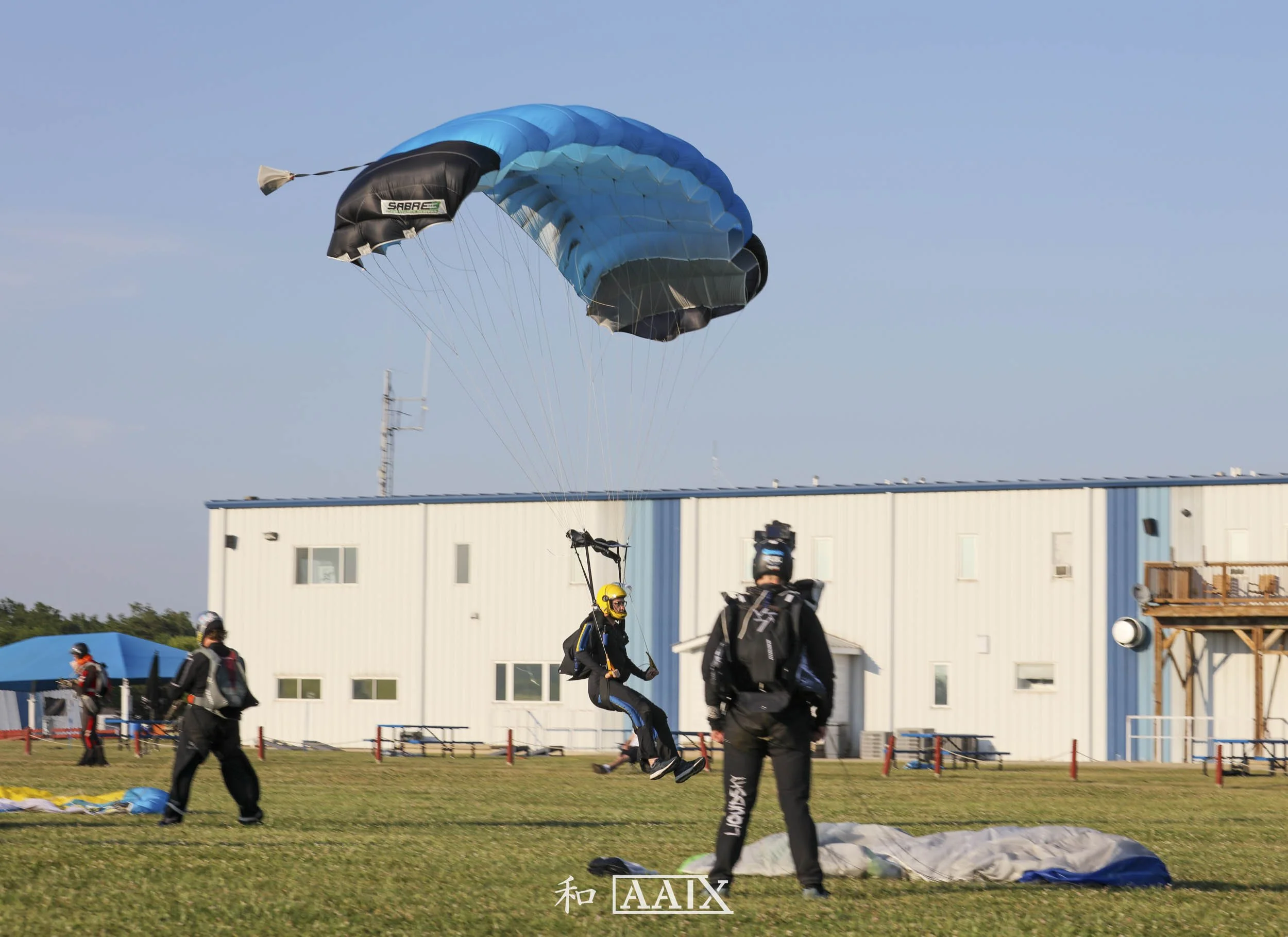 Parachutist landing on grassy field with others preparing for jump, a white building in background, and clear blue sky.