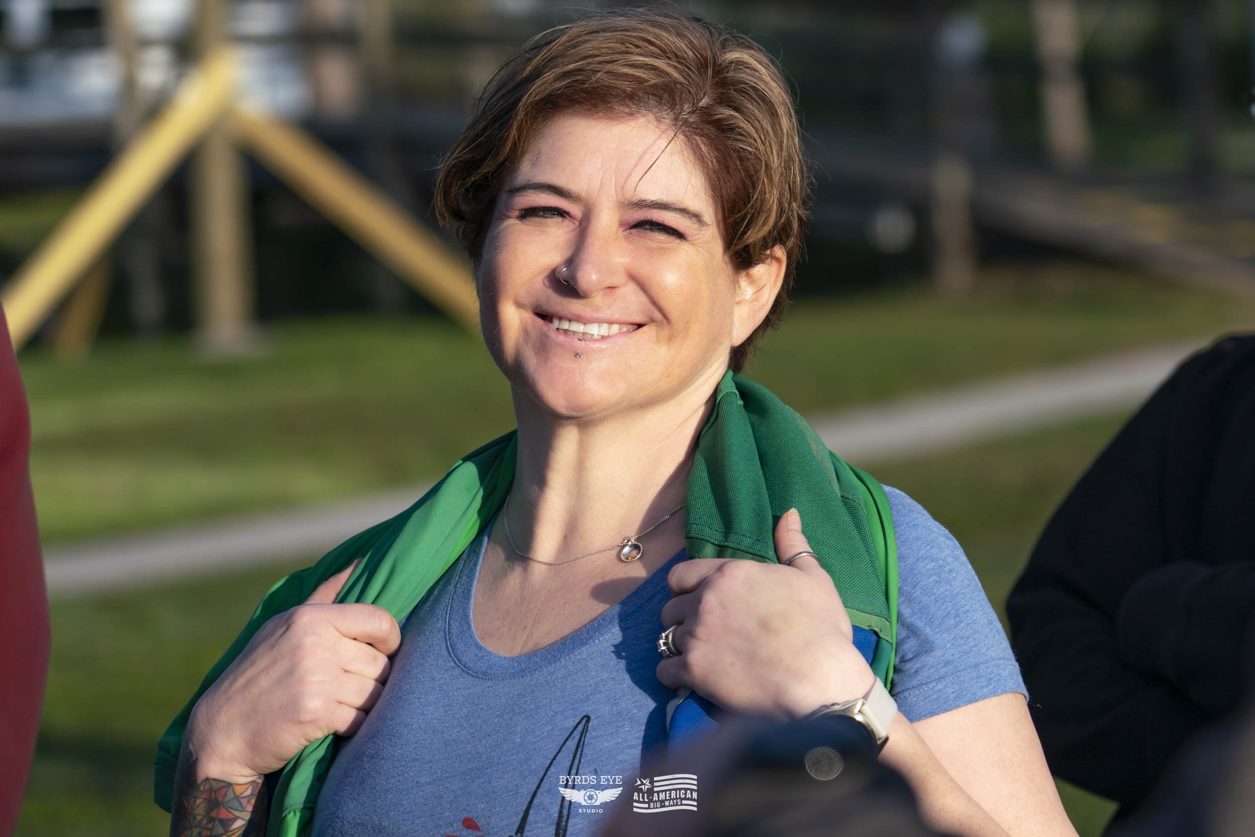 A smiling woman with short brown hair and light skin, wearing a blue T-shirt and a green backpack, standing outdoors in a park-like setting.