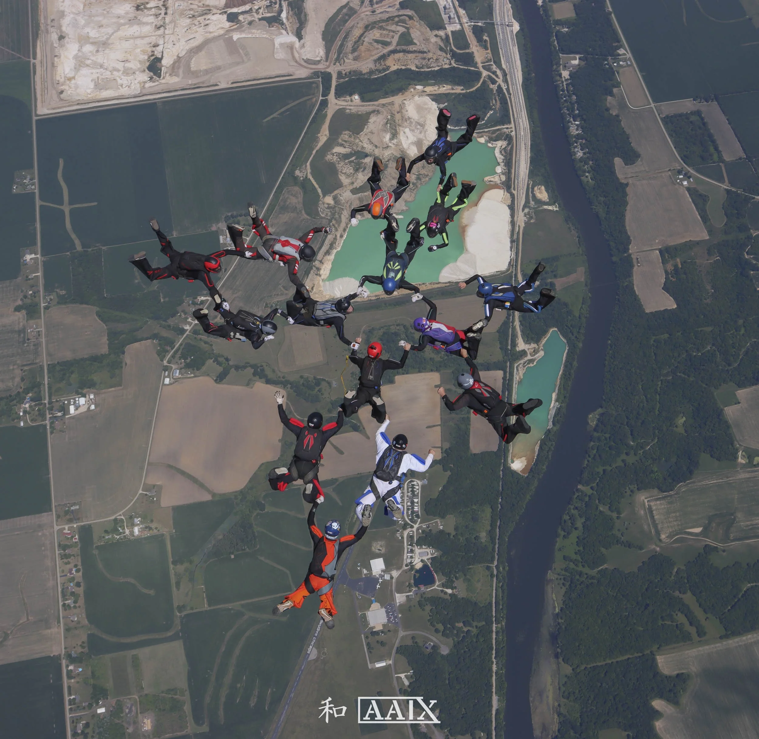 Group of skydivers forming a star shape while free-falling over farmland and water bodies.